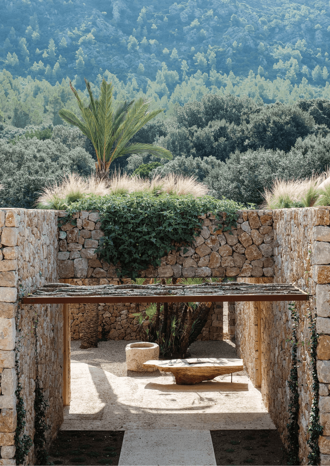 Es Racó d'Artà, Balearic Islands, Spain — Stone archway framing courtyard with palm tree, olive groves, and coniferous forest backdrop.