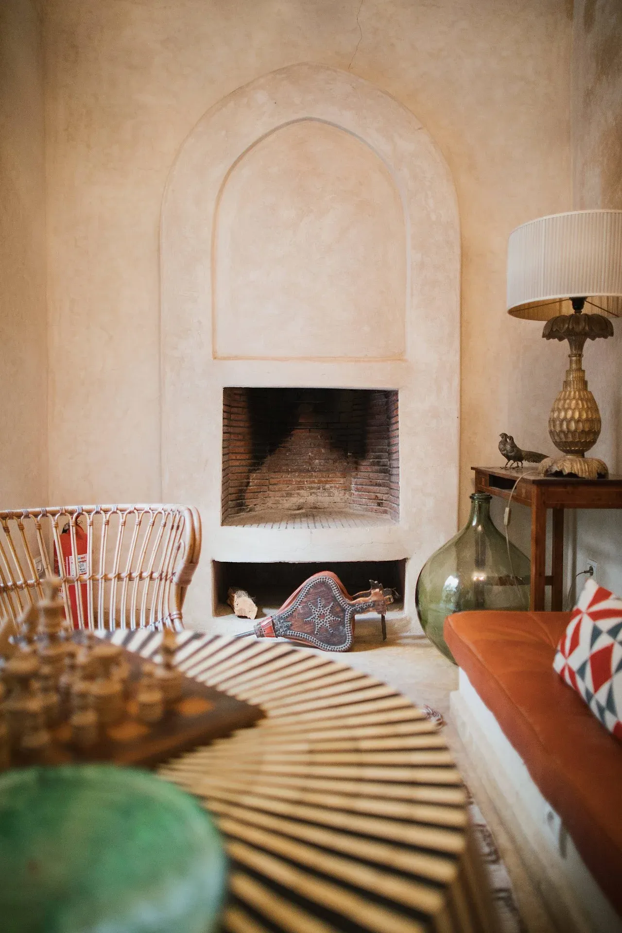 El Fenn, Africa — Arched fireplace in cream plaster with exposed brick interior, rattan chair, brass lamp, and decorative objects in minimalist Moroccan room.