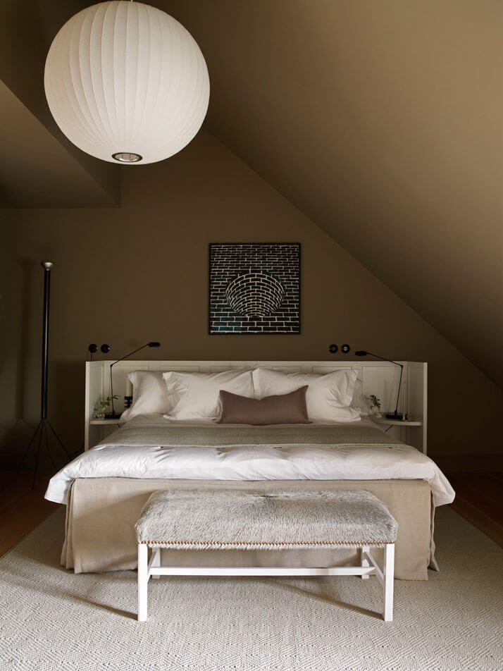 Ett Hem, Europe — Minimalist attic bedroom with linen bed, paper pendant light, brick tunnel artwork, and natural wood bench under sloped ceiling.