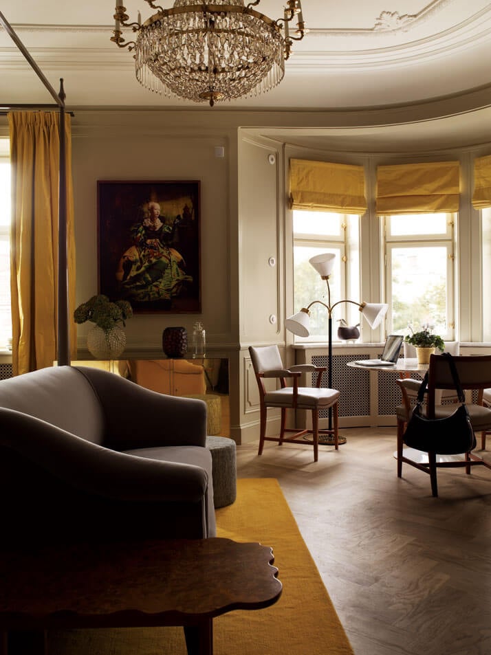 Ett Hem, Europe — Luminous sitting room with curved ceiling, brass chandelier, mustard curtains, gray curved sofa, and bay windows overlooking gardens.