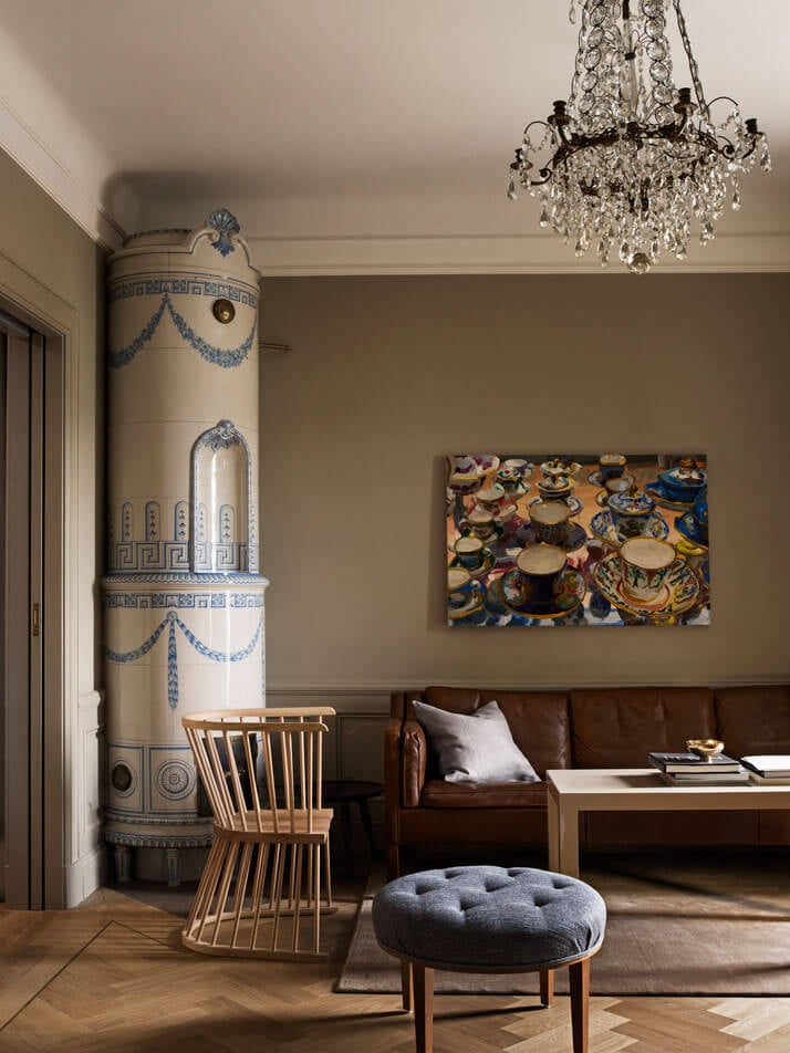 Ett Hem, Europe — Neoclassical stove, leather sofa, crystal chandelier, and contemporary ceramic artwork in refined living room with herringbone flooring.