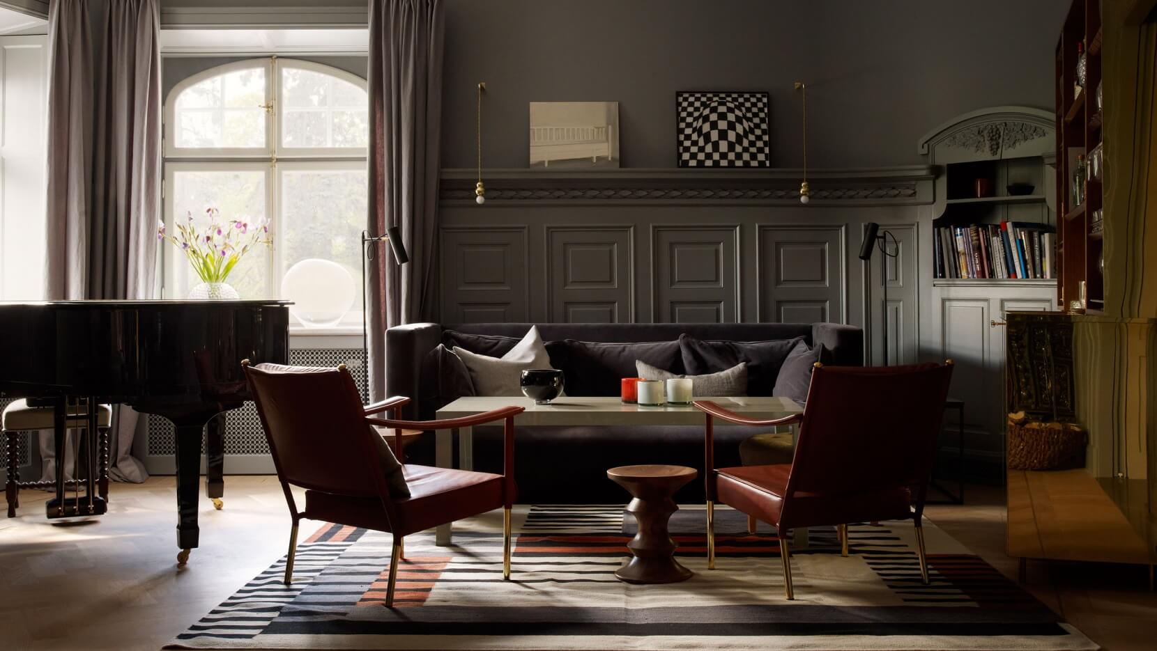 Ett Hem, Europe — Black grand piano, cognac leather chairs, and dark sofa in sunlit living room with paneled walls and geometric rug.