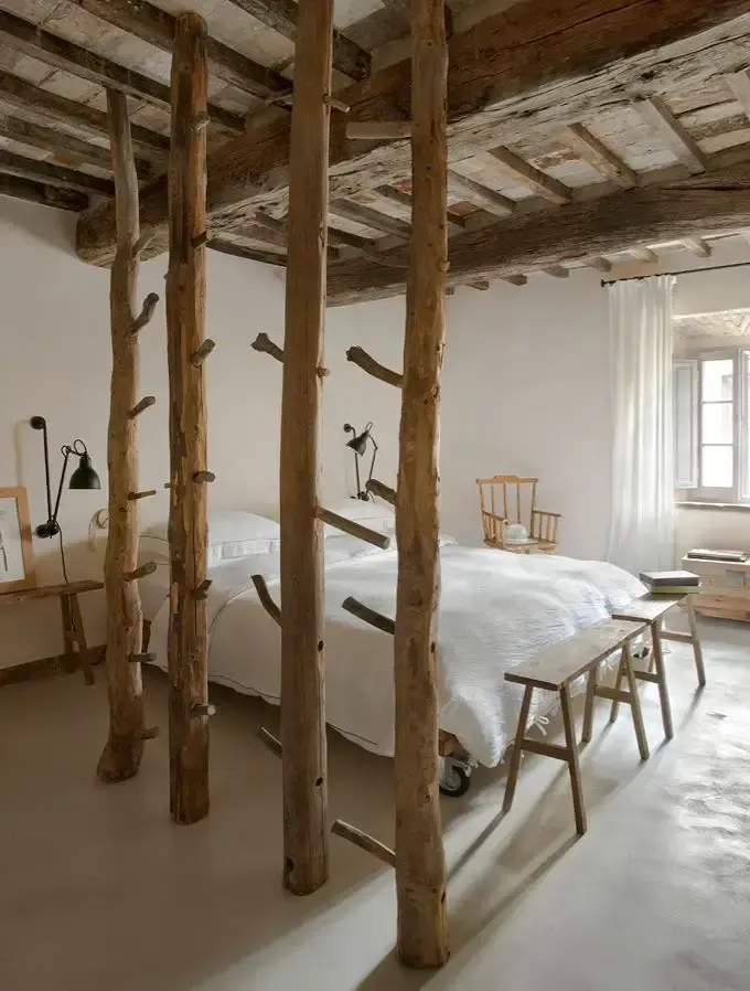 Monteverdi, Europe — Rustic canopy bed with raw timber posts and branches, white linens, exposed wooden ceiling beams, minimalist luxury bedroom design.
