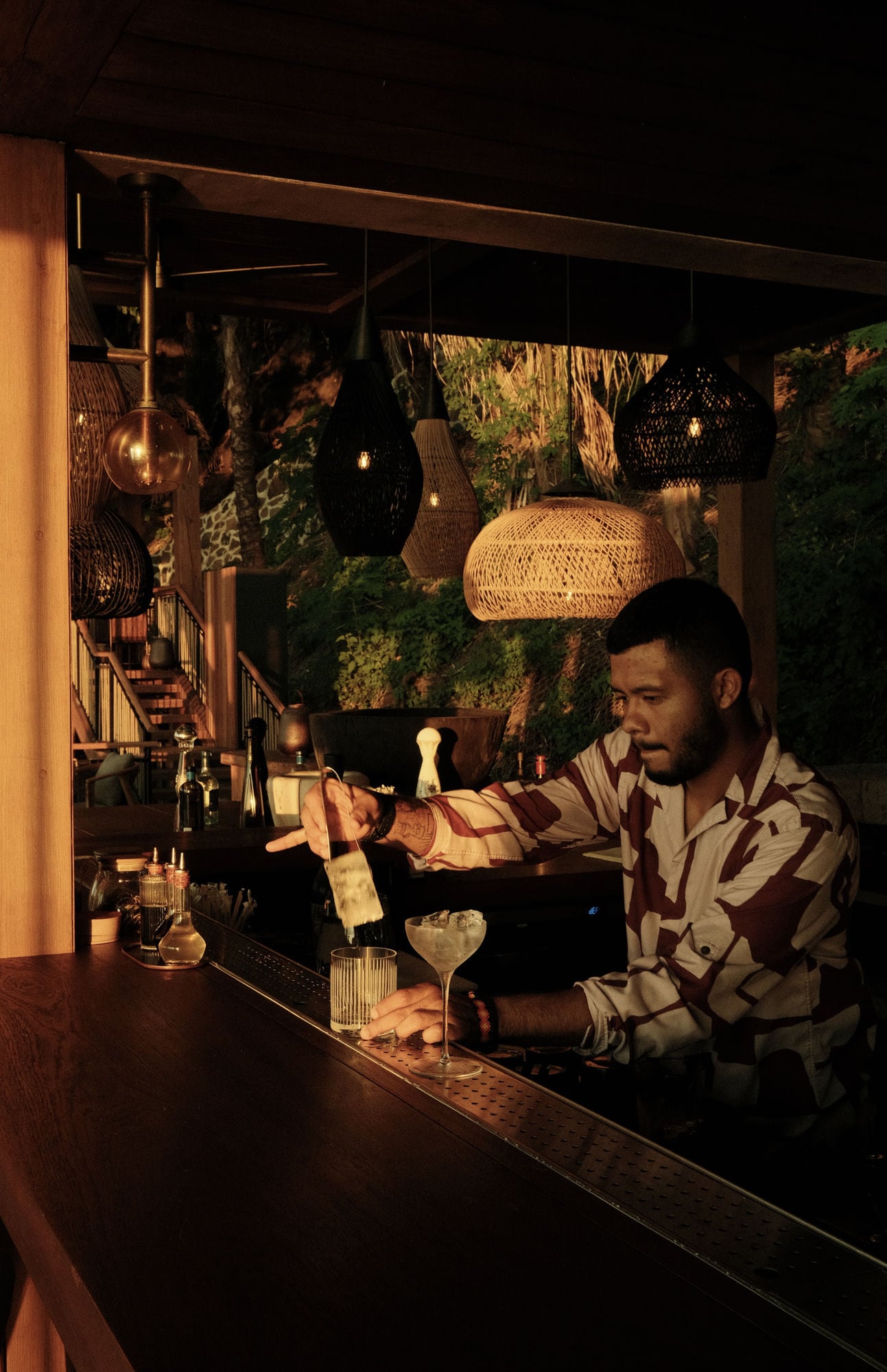 A bartender finishing a drink at a timber bar in the evening, woven lanterns and dense greenery behind