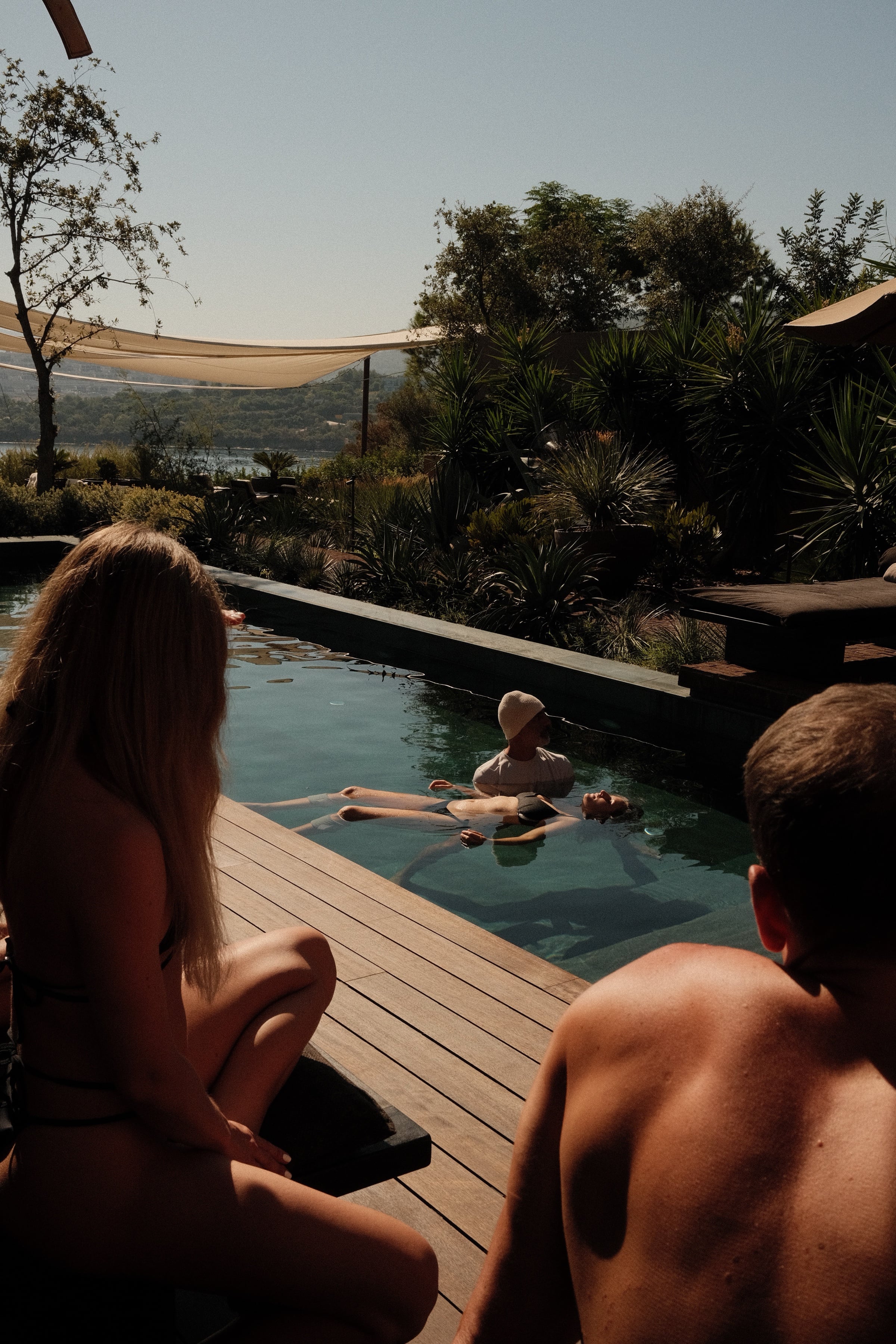 Guests at the poolside in late afternoon light, one floating in the water supported by a practitioner, palms and open water beyond