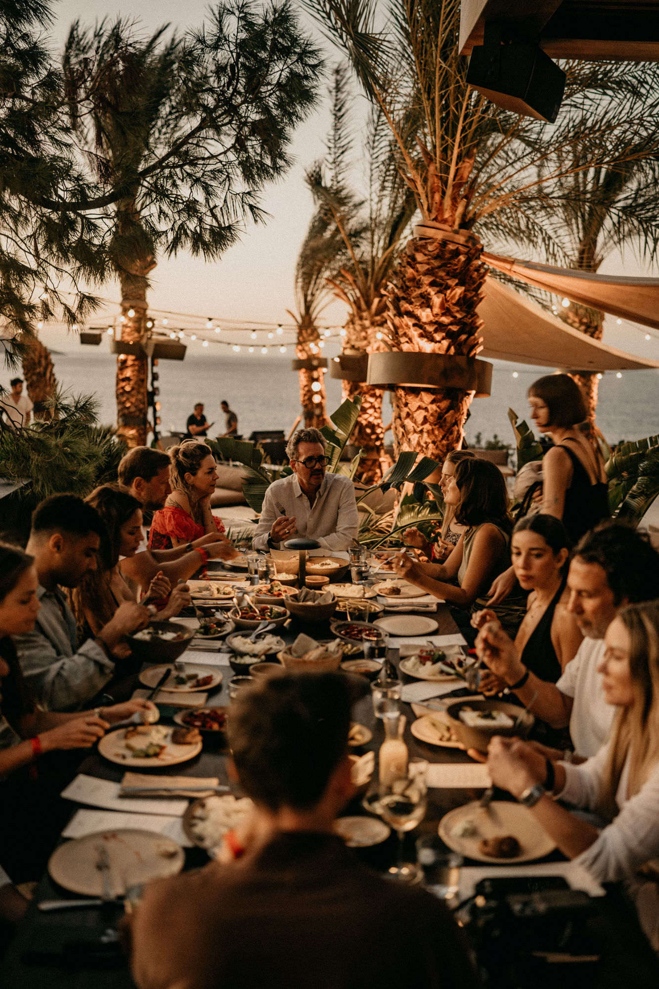 A long shared table at sunset beneath palms and string lights, guests dining together beside the sea