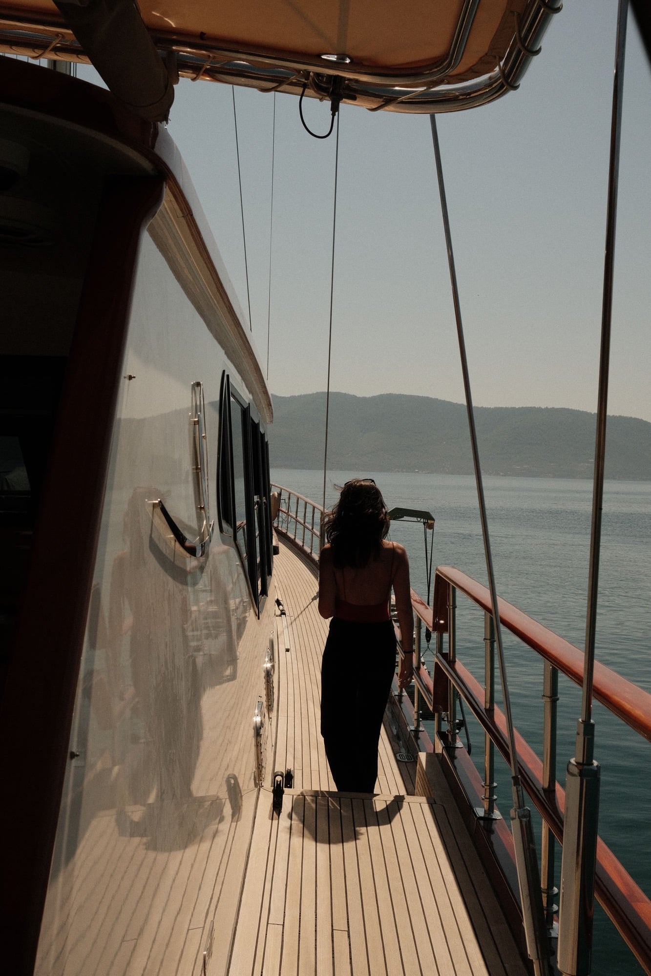 A guest walking the teak side deck of a wooden yacht at sea, hills on the horizon