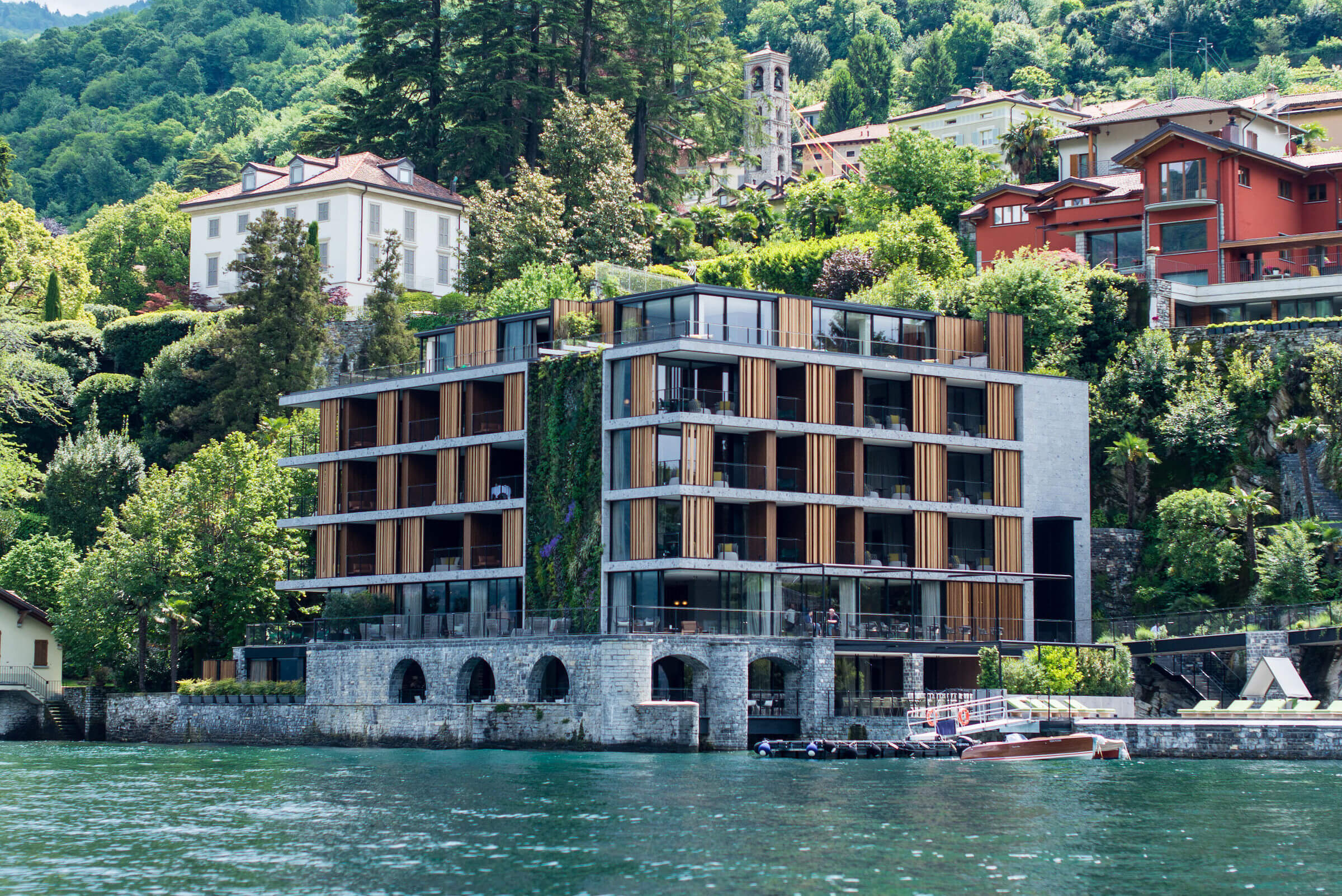 il Sereno Lago di Como, Lake Como, Italy — Modern waterfront hotel with wood-and-glass facade, stone arcade, terraced balconies overlooking alpine lake and hillside village architecture.