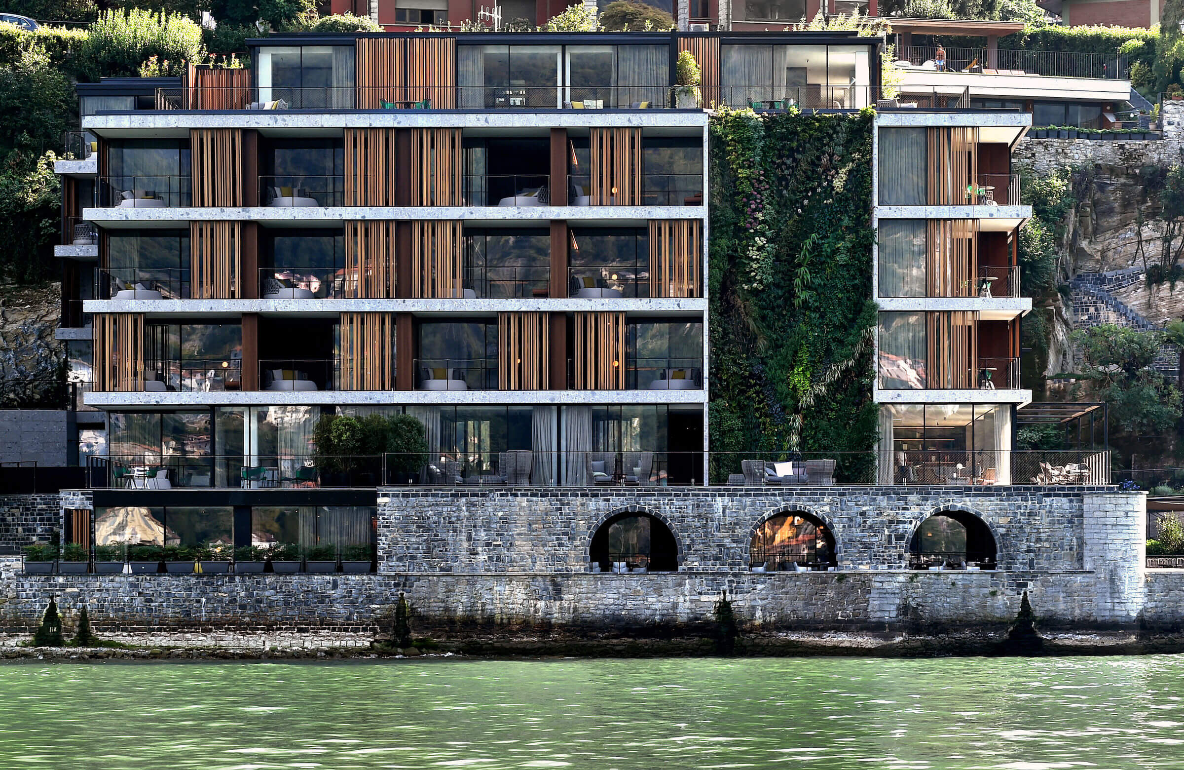il Sereno Lago di Como, Lake Como, Italy — Waterfront modernist facade with stacked timber and glass panels, arched stone loggia, contemporary luxury architecture on Alpine lakeside.