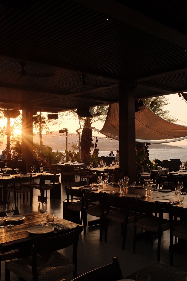 Scorpios Bodrum, Turkey — Waterfront dining pavilion at sunset with wooden tables, canvas sail canopies, and Aegean Sea views through open timber architecture.