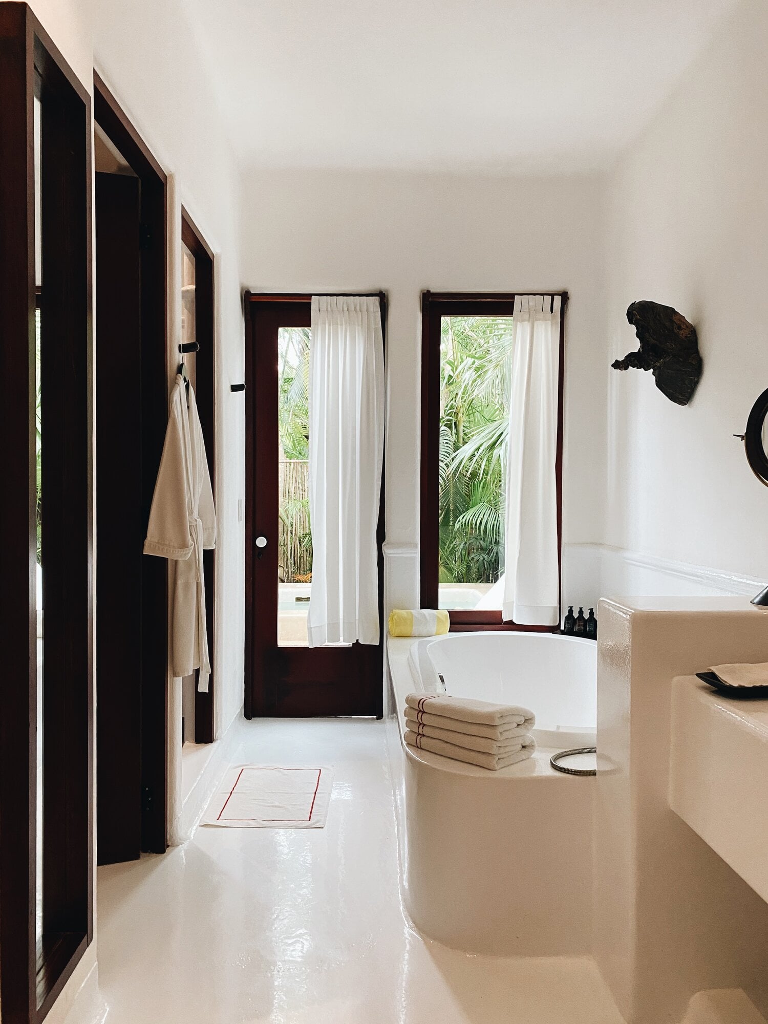 Hotel Esencia, Mexico — Minimalist bathroom with dark wood-framed doors, white marble flooring, soaking tub, and tropical garden views through sheer curtains.