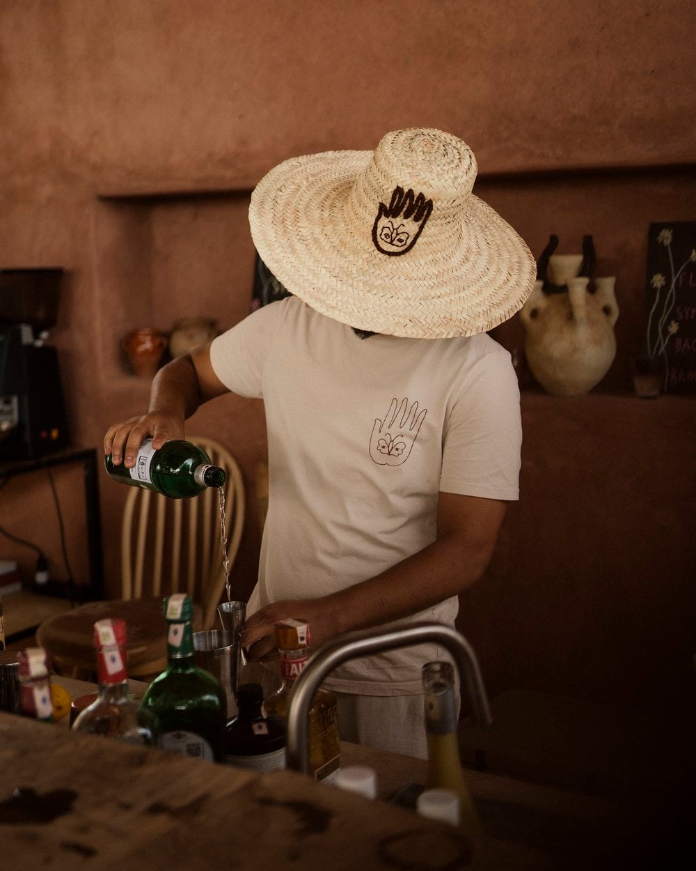 Farasha Farmhouse, Africa — Person in cream linen pouring wine at terracotta-walled bar, wearing straw sombrero with hand motif, warm golden light, rustic hospitality aesthetic.