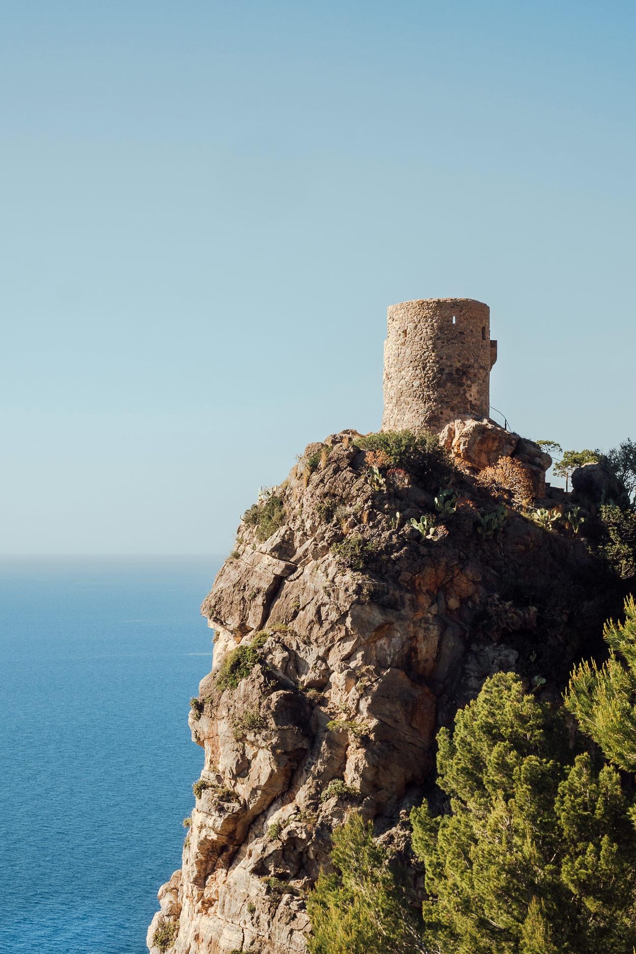 Hideaway, Balearic Islands, Spain — Weathered stone watchtower perched on rocky coastal cliff overlooking azure Mediterranean, sparse vegetation and clear sky.