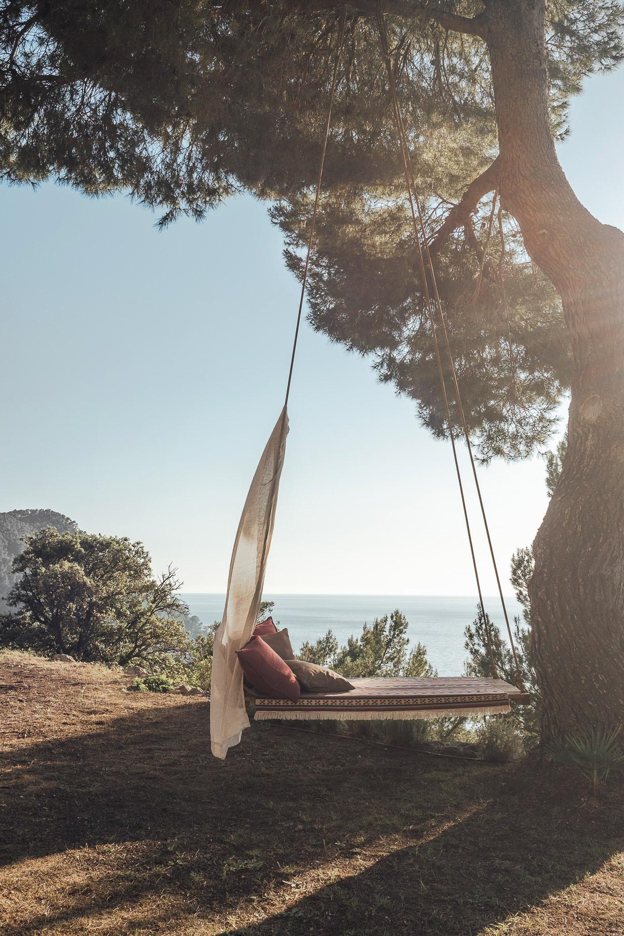 Hideaway, Balearic Islands — Suspended wooden daybed with linen drape beneath pine trees overlooking Mediterranean sea, dappled sunlight, coastal retreat.