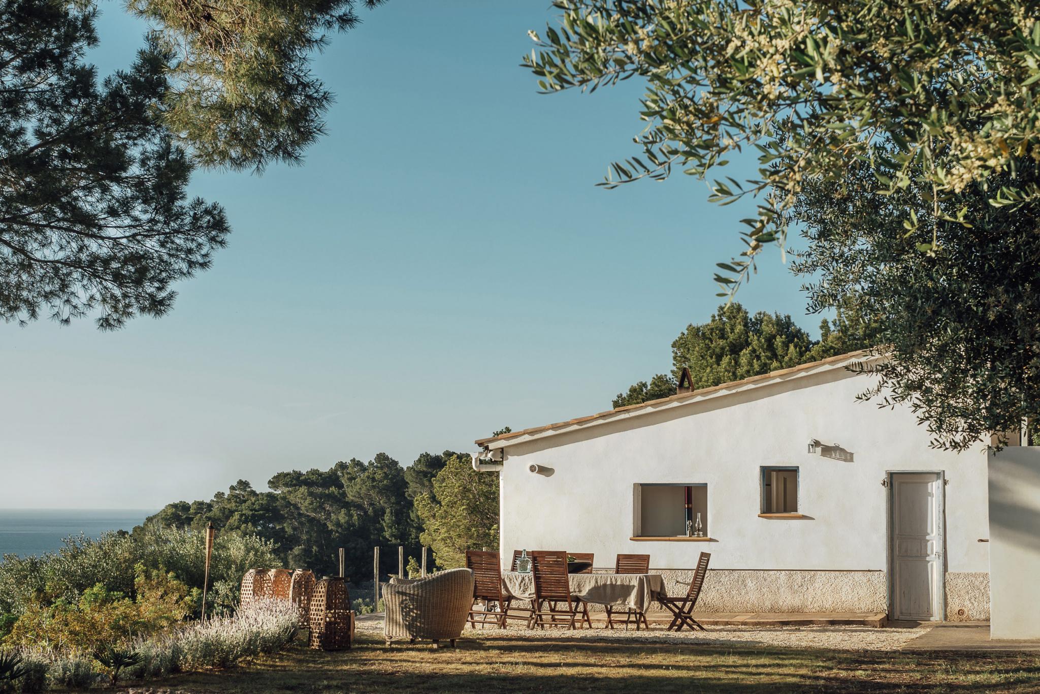 Hideaway, Balearic Islands, Spain — Minimalist whitewashed villa with wooden chairs on terrace overlooking Mediterranean sea and pine forest under clear sky.