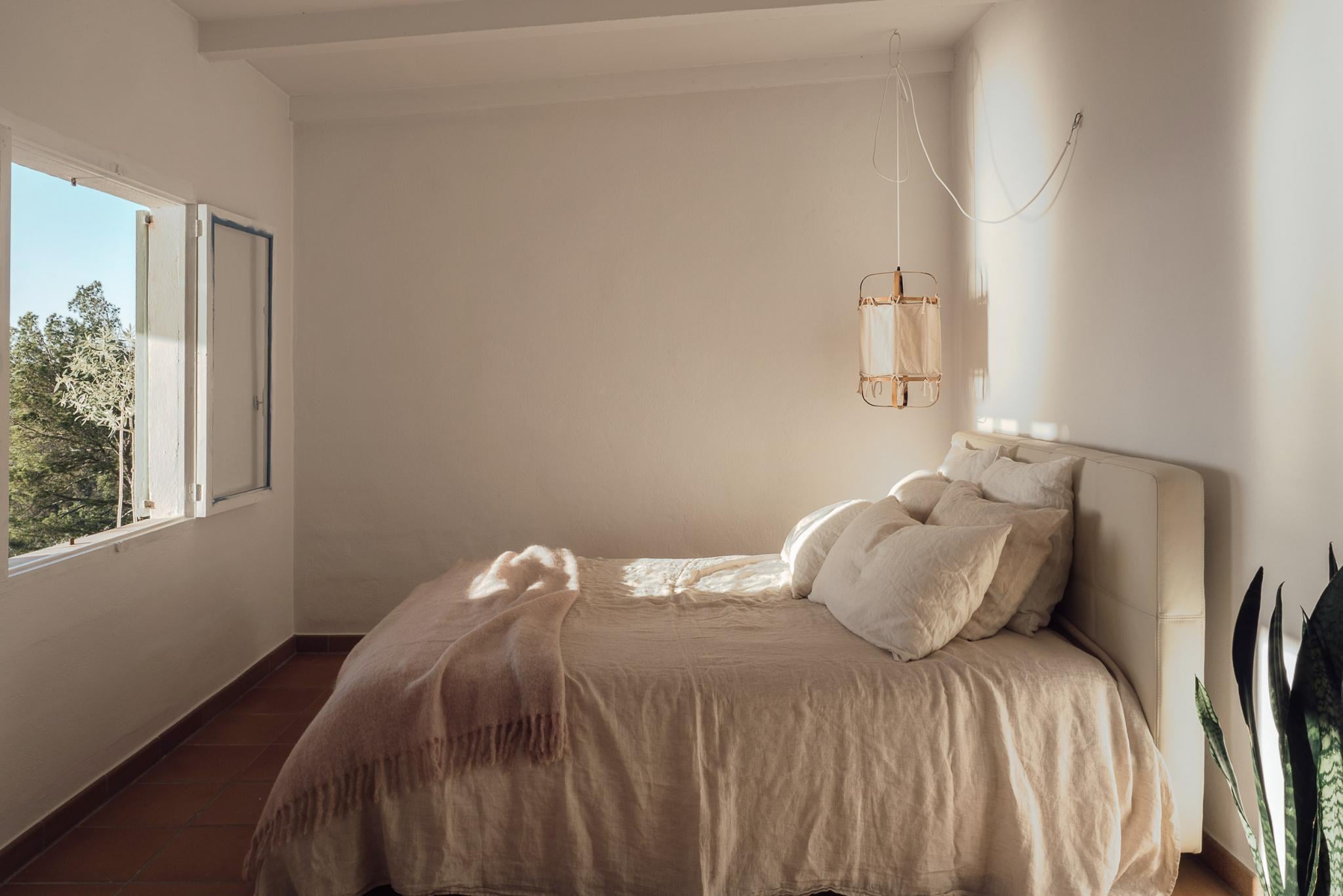 Hideaway, Balearic Islands, Spain — Minimalist bedroom with natural linen bedding, wooden pendant light, terracotta floor, and sunlit window overlooking Mediterranean landscape.