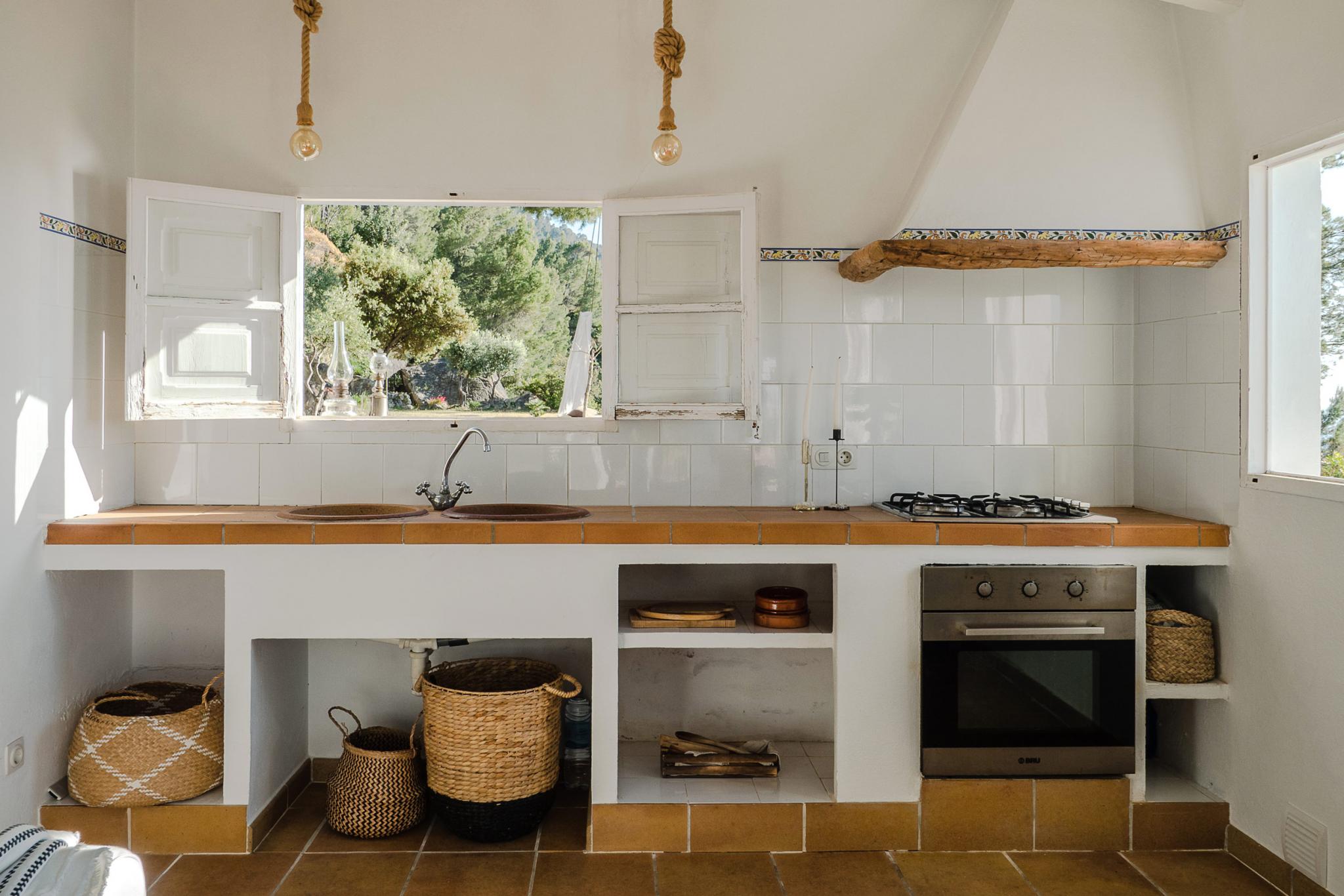 Hideaway, Balearic Islands — Minimalist open kitchen with terracotta tile counters, white subway tile backsplash, woven storage baskets, and expansive windows overlooking Mediterranean landscape.