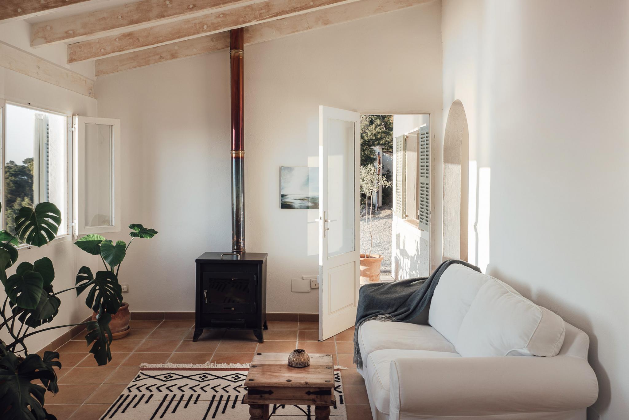 Hideaway, Balearic Islands, Spain — Minimalist living room with exposed wooden beams, white walls, black wood stove, cream linen sofa, terracotta tile flooring, and view to Mediterranean courtyard.