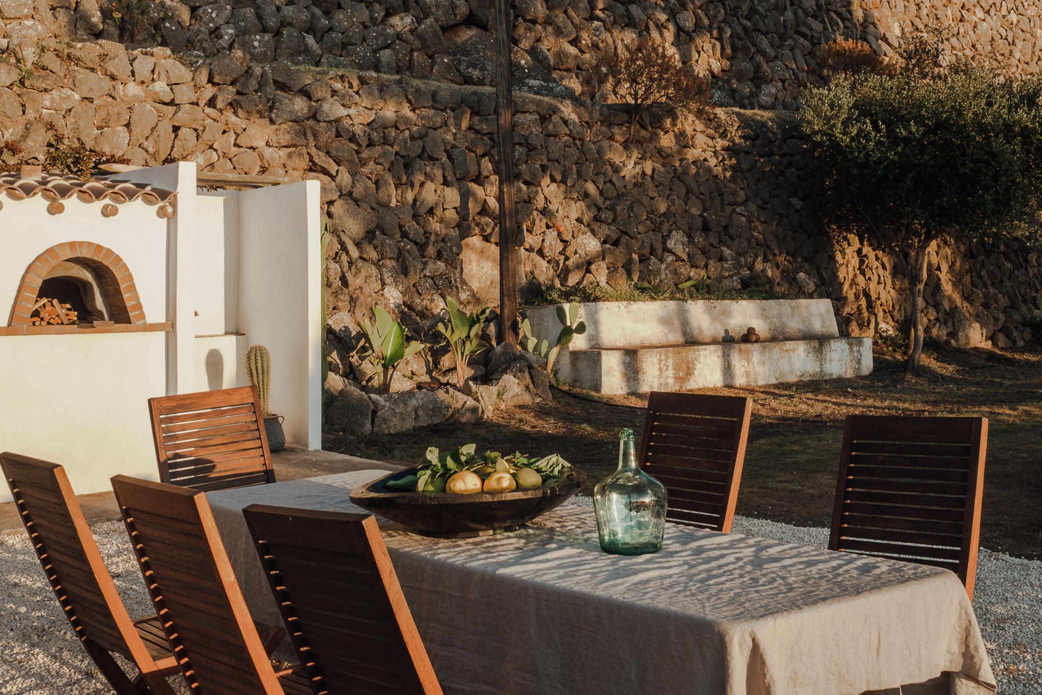 Hideaway, Balearic Islands, Spain — Rustic stone courtyard with wooden dining chairs, linen-draped table, ceramic bowl of citrus, glass carafe, and whitewashed building with brick oven.