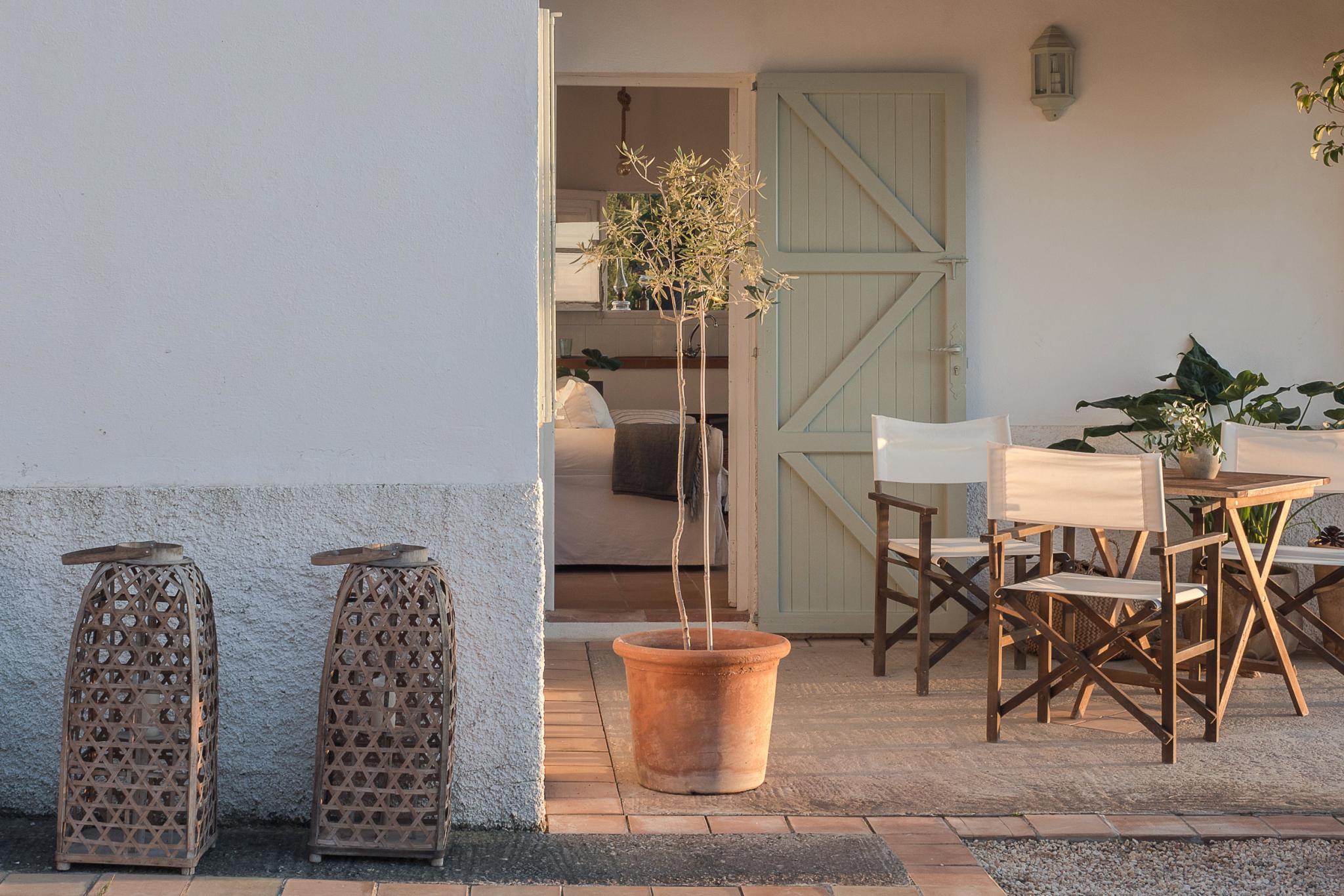 Hideaway, Balearic Islands — Whitewashed patio with woven lanterns, terracotta planter, wooden director's chairs, and sage-green barn door opening to bedroom.
