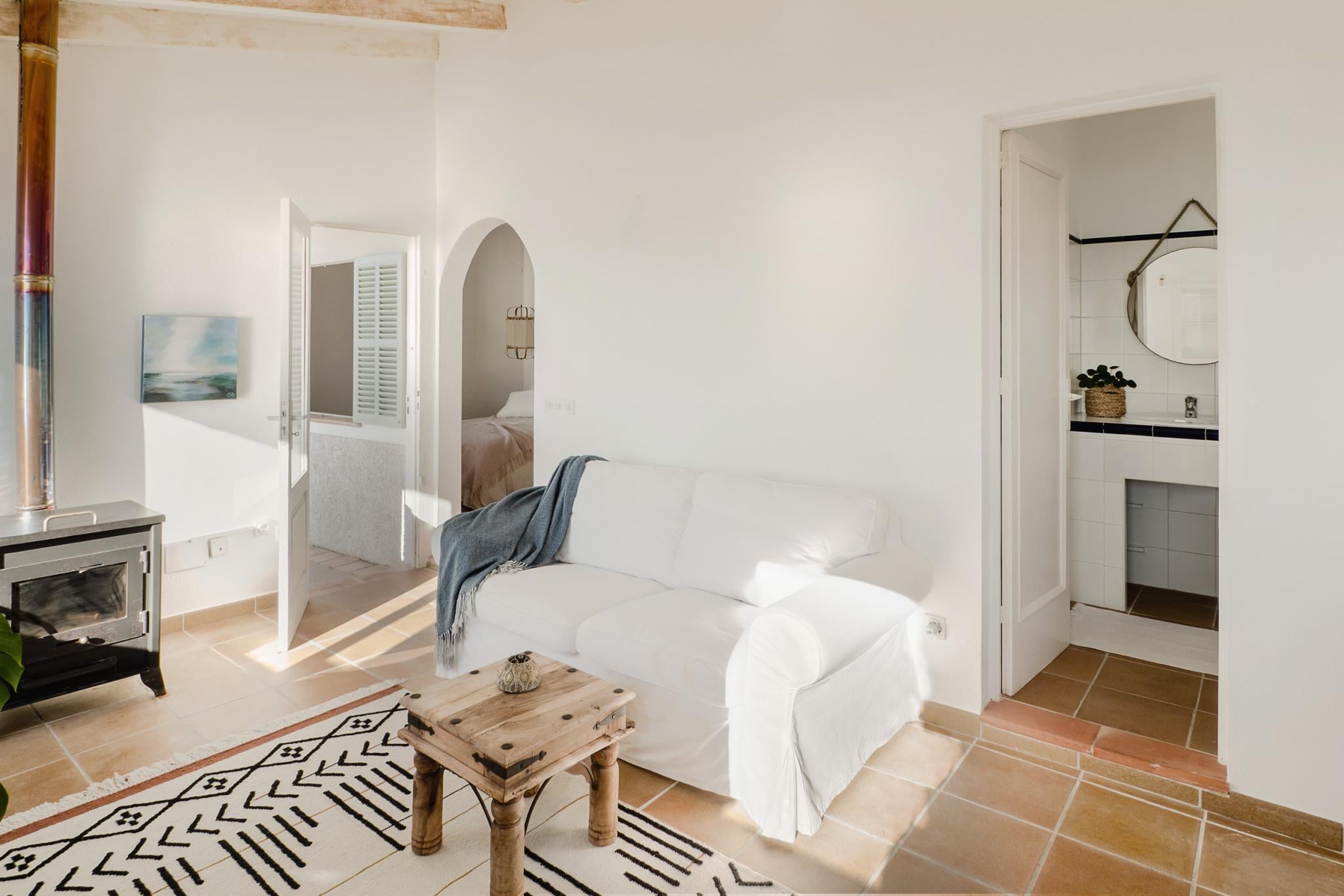 Hideaway, Balearic Islands — Bright living room with white linen sofa, terracotta tile floors, wooden beams, arched doorway to bedroom, and minimalist Mediterranean design.