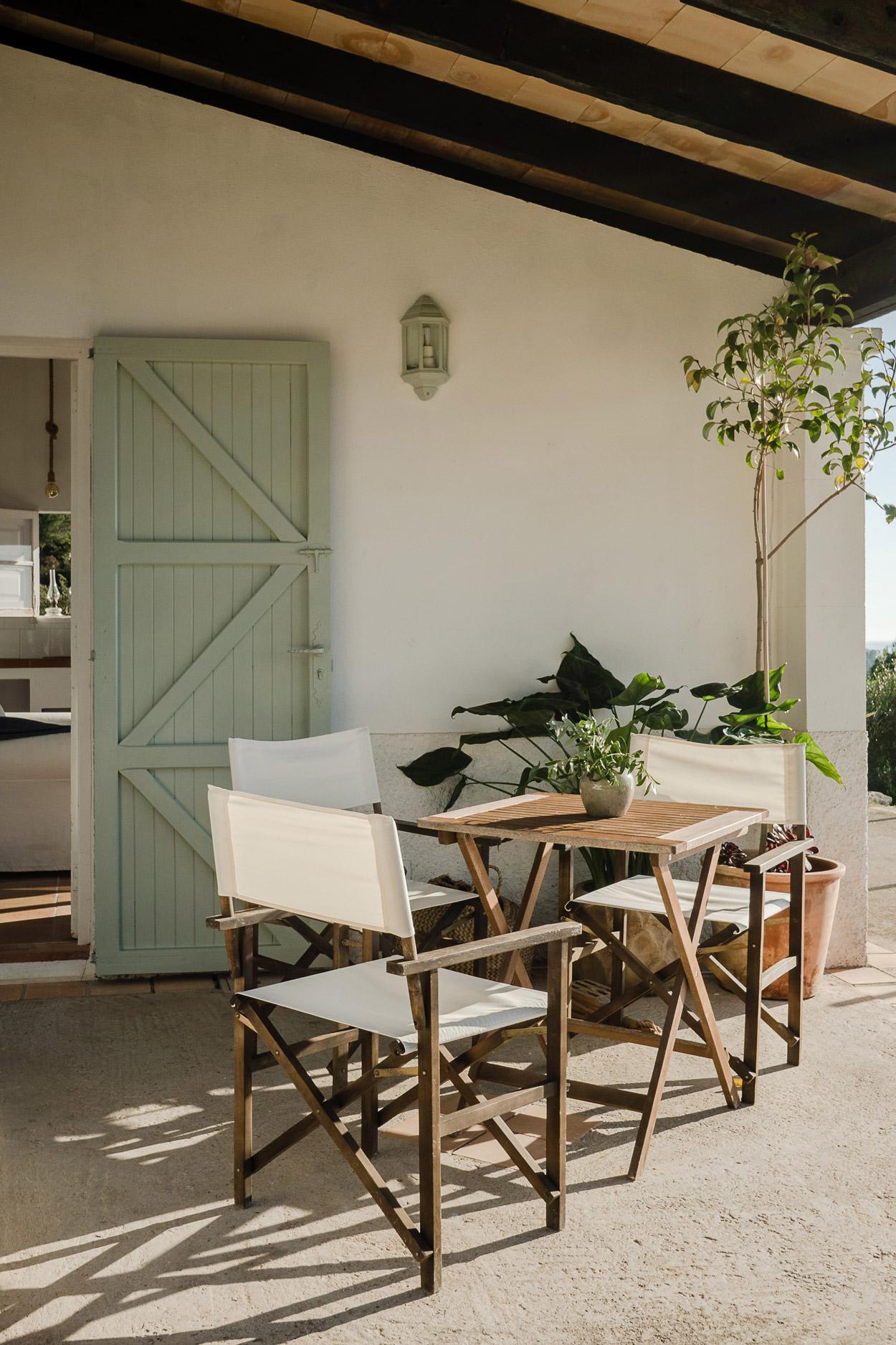 Hideaway, Balearic Islands — Shaded terrace with wooden director's chairs, natural linen cushions, and potted plants beside sage-green barn door and white-walled pavilion.