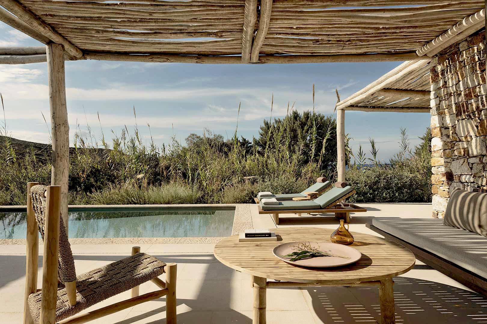 The Rooster, Europe — Wooden pergola shading a terrace with loungers, round dining table, and pool overlooking Mediterranean landscape with olive trees and scrubland.