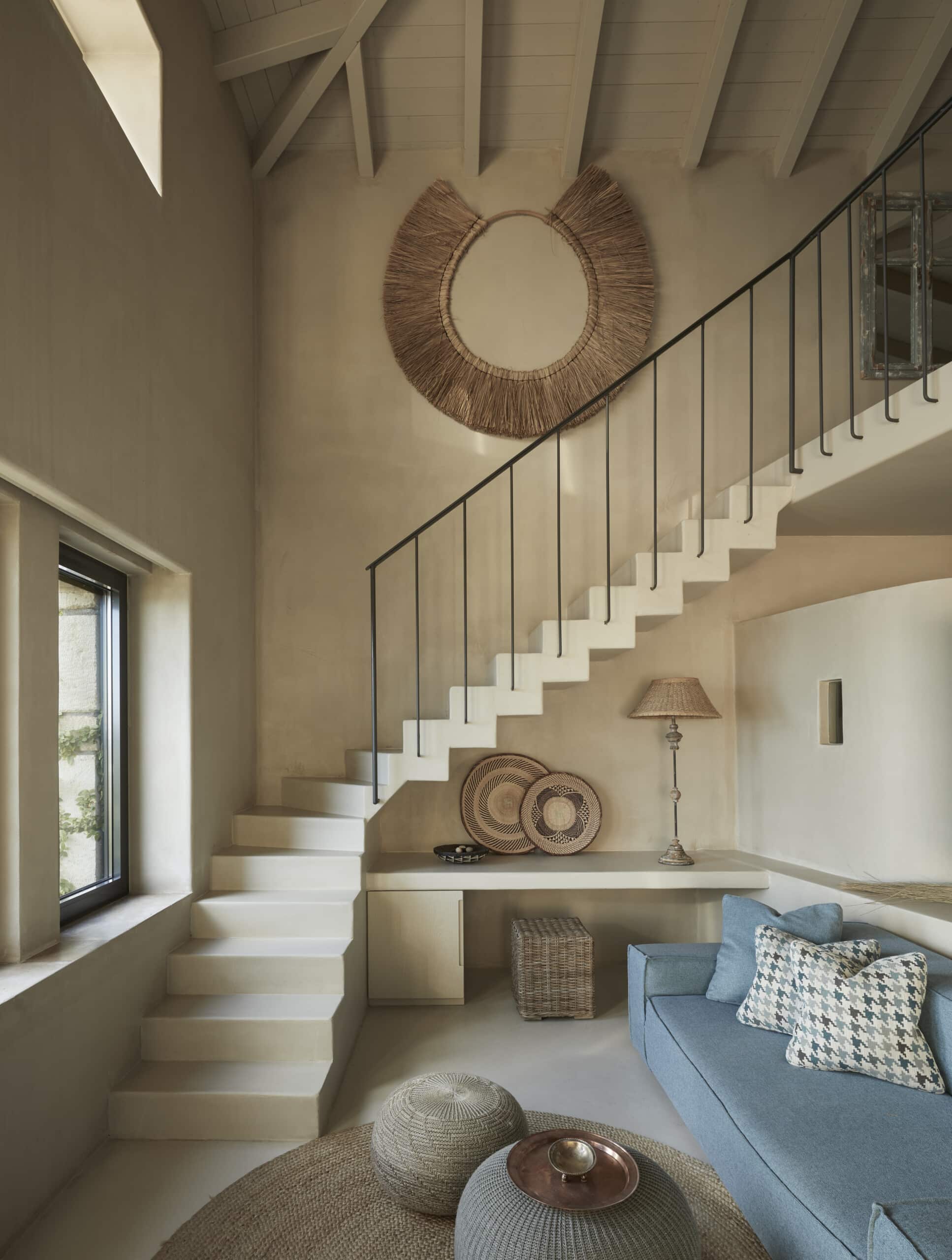 F Zeen Retreat, Europe — Double-height stairwell with woven mirror and rattan accents, cream walls, natural light, minimalist design with blue seating nook.
