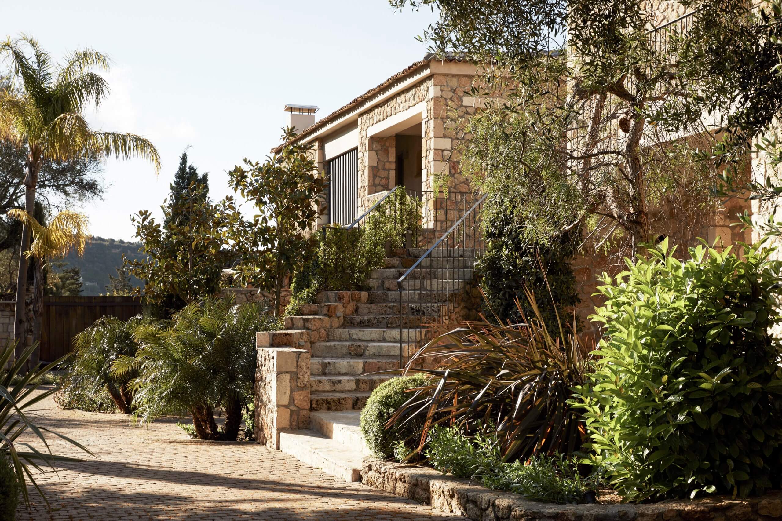 F Zeen Retreat, Europe — Stone villa entrance with metal staircase, surrounded by Mediterranean plants and palm trees, warm natural light casting shadows on brick courtyard.