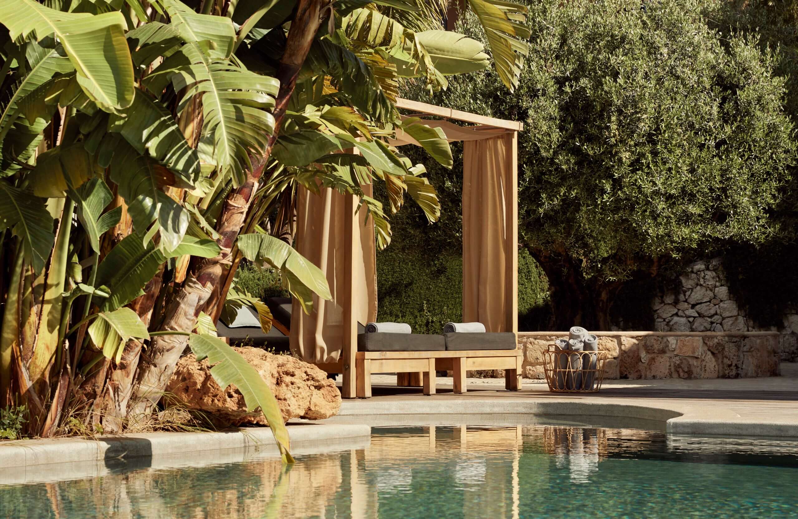 F Zeen Retreat, Europe — Minimalist wooden pergola with draped curtains beside pool, surrounded by palm and olive trees, stone terrace, afternoon light.