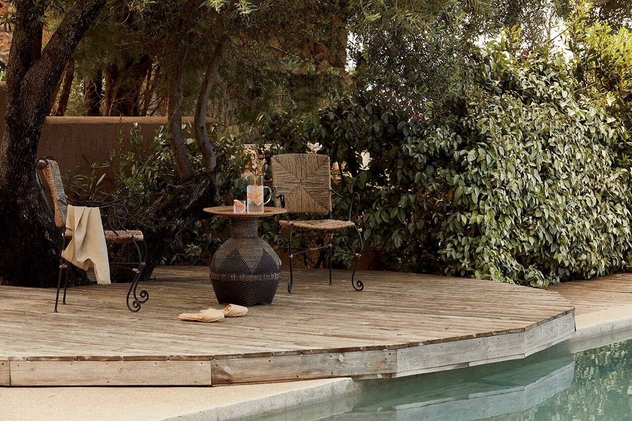 F Zeen Retreat, Europe — Curved wooden deck with woven sphere side table, iron chairs, and lush vegetation creating intimate poolside lounge setting.