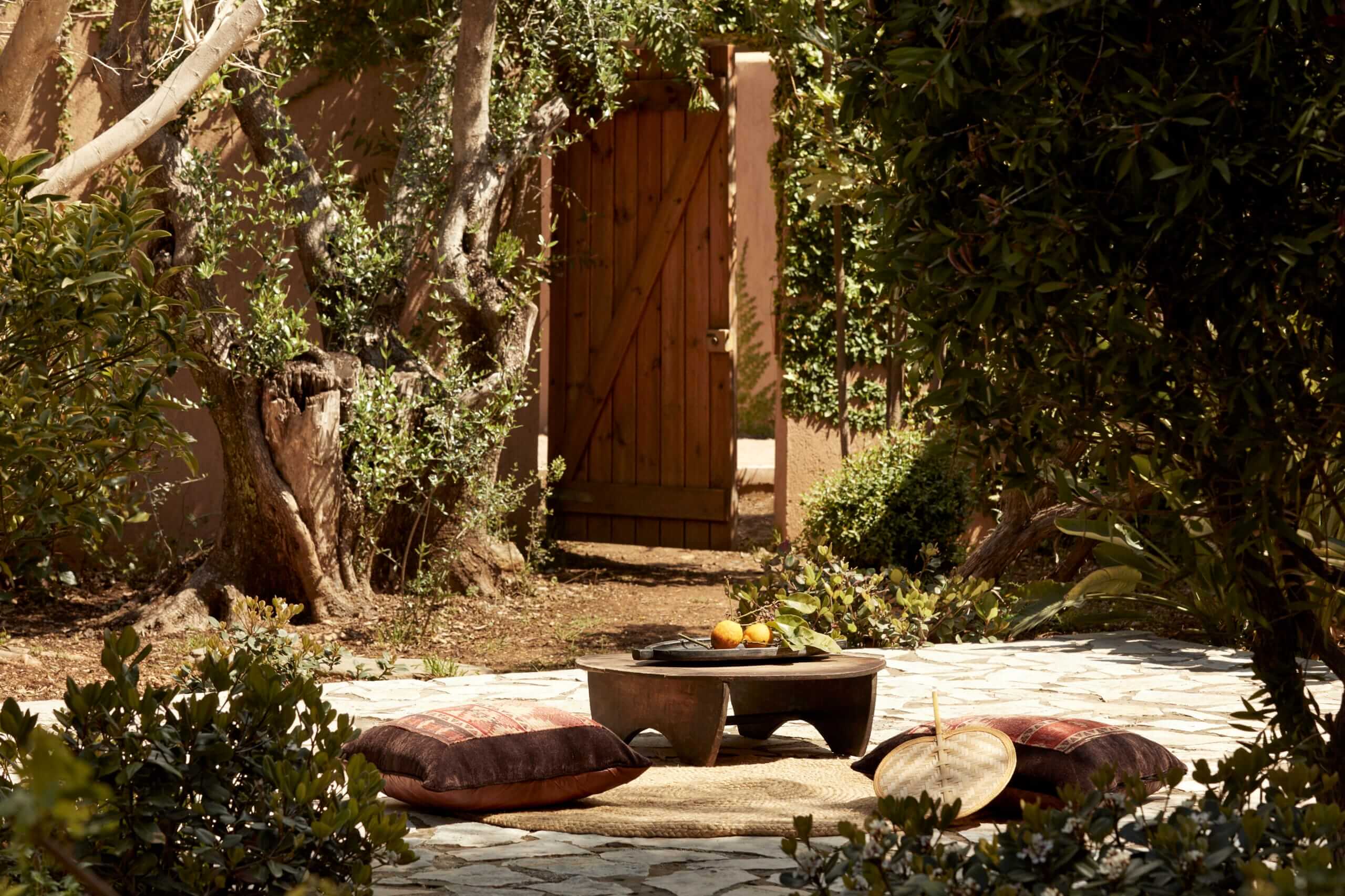 F Zeen Retreat, Europe — Shaded courtyard with stone terrace, clay vessels, woven cushions, and wooden door framed by olive trees and climbing vines.