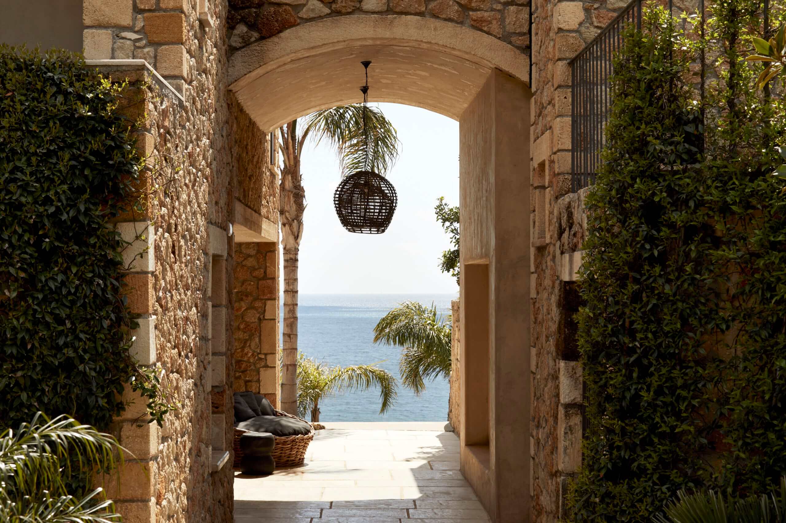 F Zeen Retreat, Europe — Stone archway with woven pendant light framing Mediterranean sea view, climbing ivy, palm fronds, and sunlit terrace.
