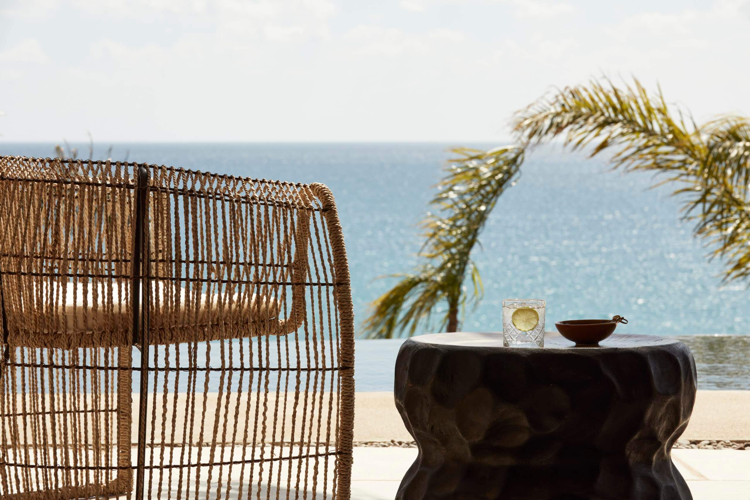 F Zeen Retreat, Europe — Woven rattan chair and sculptural black table with glass of water overlooking turquoise sea and palm fronds, luxury coastal resort design.