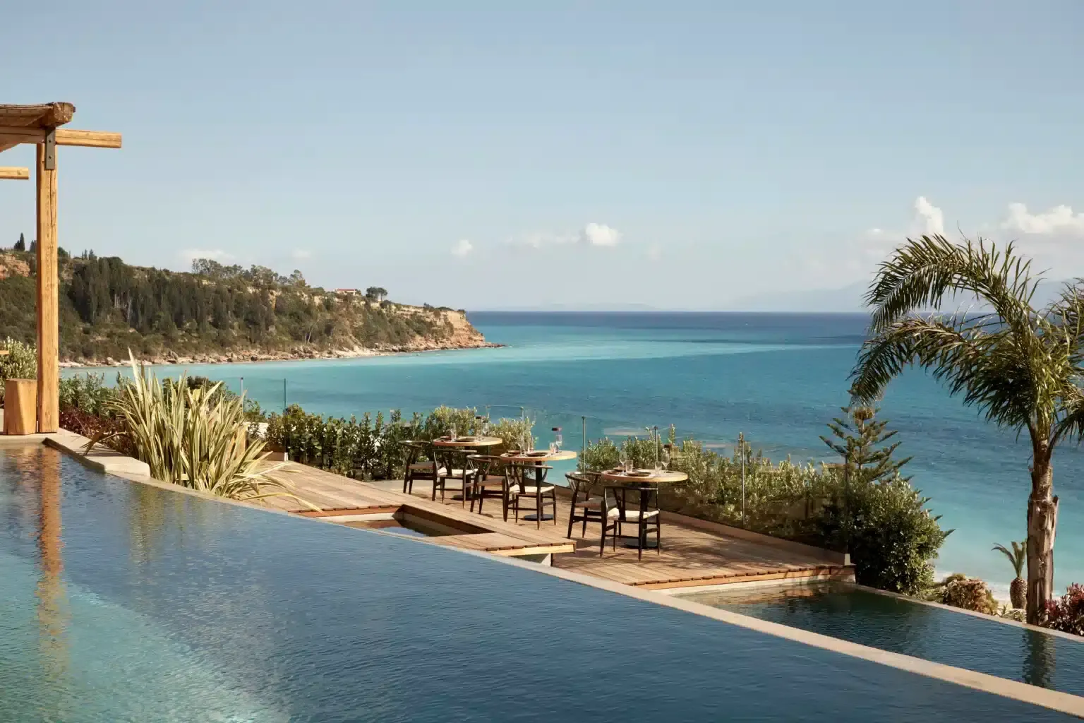 F Zeen Retreat, Europe — Infinity pool overlooking turquoise Mediterranean cove with wooden terrace, palm trees, and clifftop village nestled in pine forest.