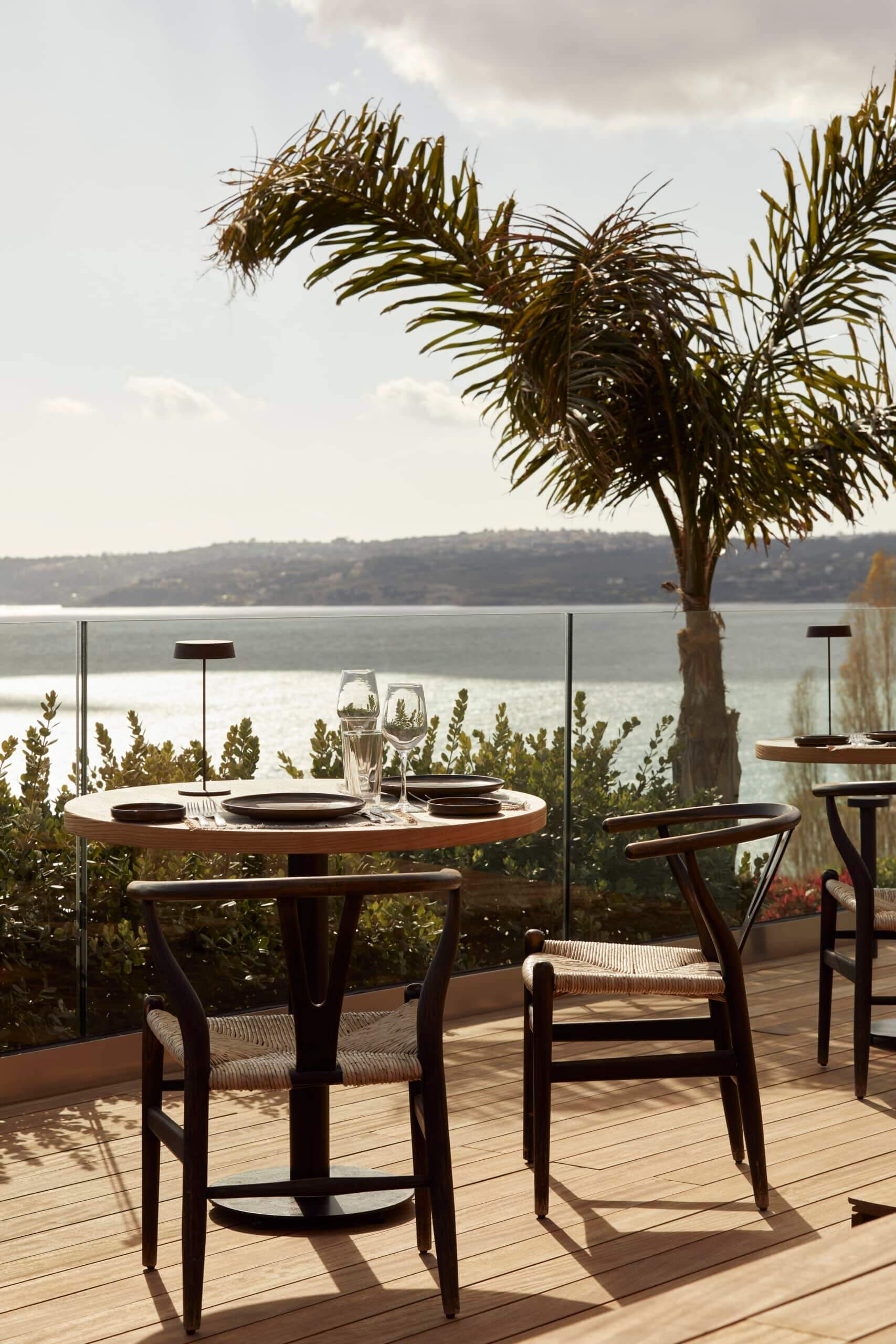 Selini, Europe — Waterfront dining terrace with wooden chairs, palm tree, and panoramic bay views at sunset.
