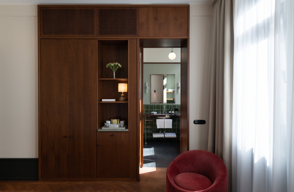 Château Royal, Europe — Walnut wardrobe with open shelving, pendant light, and view through open door to ensuite bathroom with green tile, beside burgundy modern chair and sheer curtains.