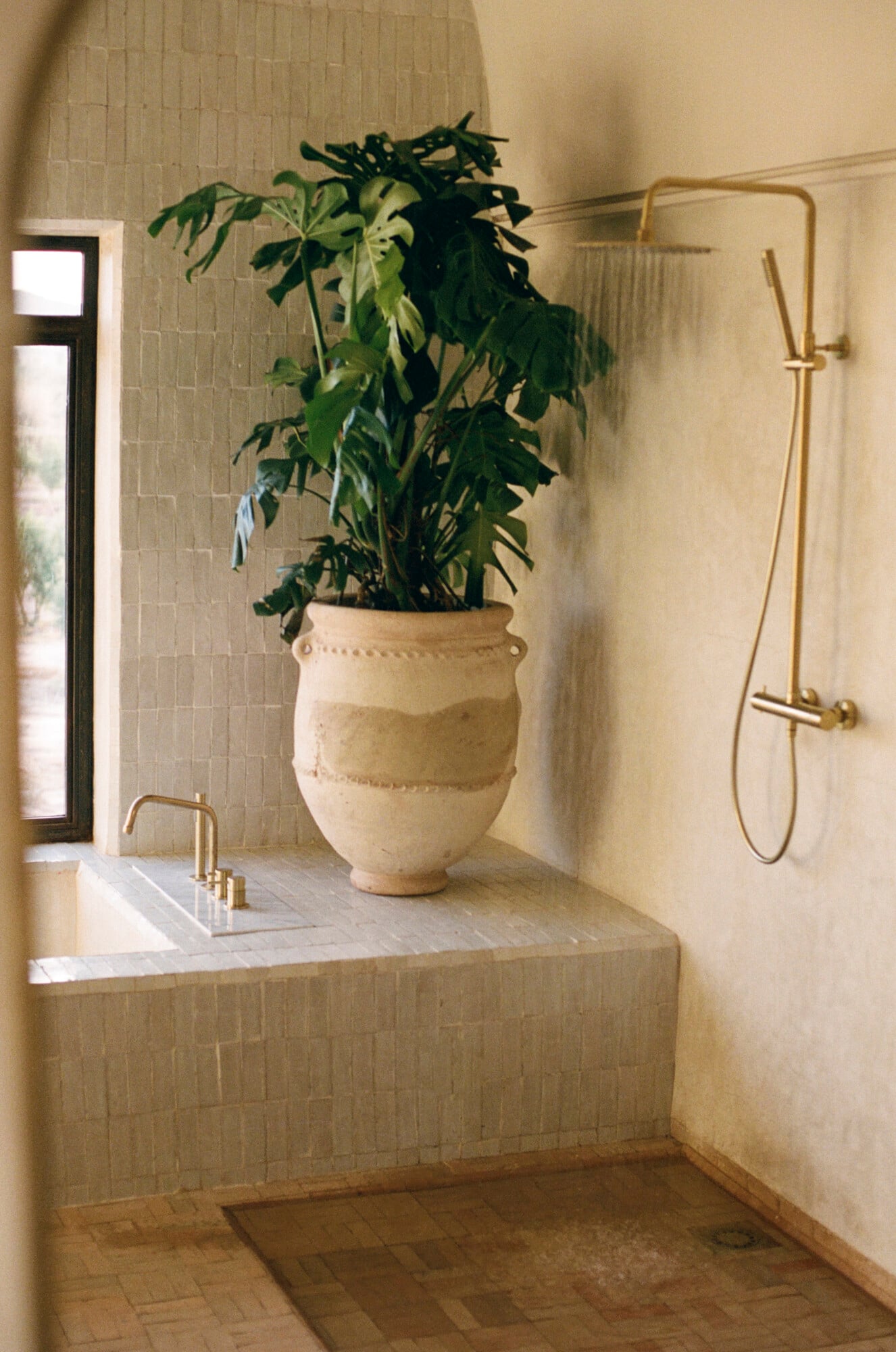 Farasha Farmhouse, Africa — Minimalist bathroom with cream tile walls, sunken soaking tub, brass fixtures, terracotta floors, and sculptural ceramic planter with trailing greenery.