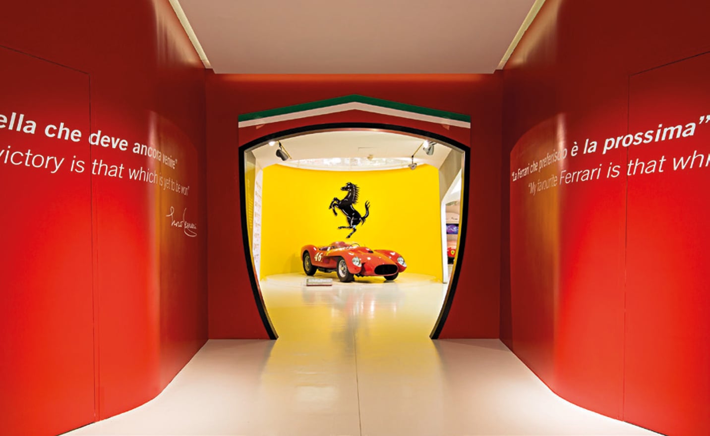 Motor Valley, Europe — Red gallery corridor with shield-shaped Ferrari showroom entrance displaying vintage red racing car beneath prancing horse logo on yellow wall.