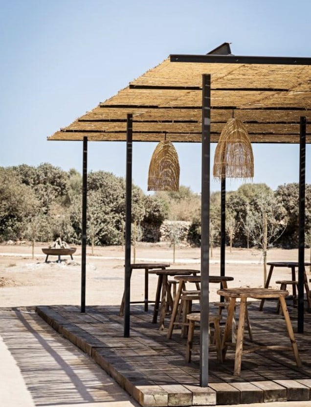 Fontenille Santa Ponsa, Balearic Islands — Wooden pergola with woven pendant lights, wooden seating, and fire pit in arid Mediterranean landscape.