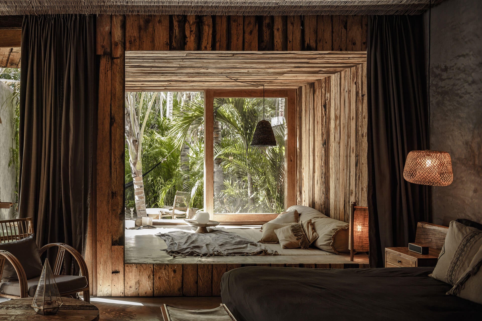 Be Tulum, Mexico — Tropical bedroom with recessed platform bed, dark linen, wooden frame windows overlooking palm garden, woven pendant lights, earth-tone curtains, concrete walls.