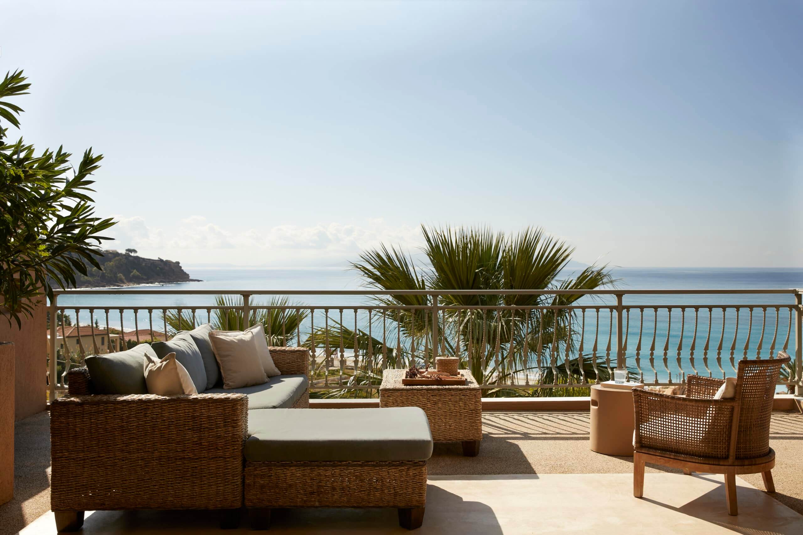 F Zeen Retreat, Europe — Coastal terrace with woven rattan seating, pale linen cushions, potted palms, and metal railings overlooking turquoise Mediterranean waters.