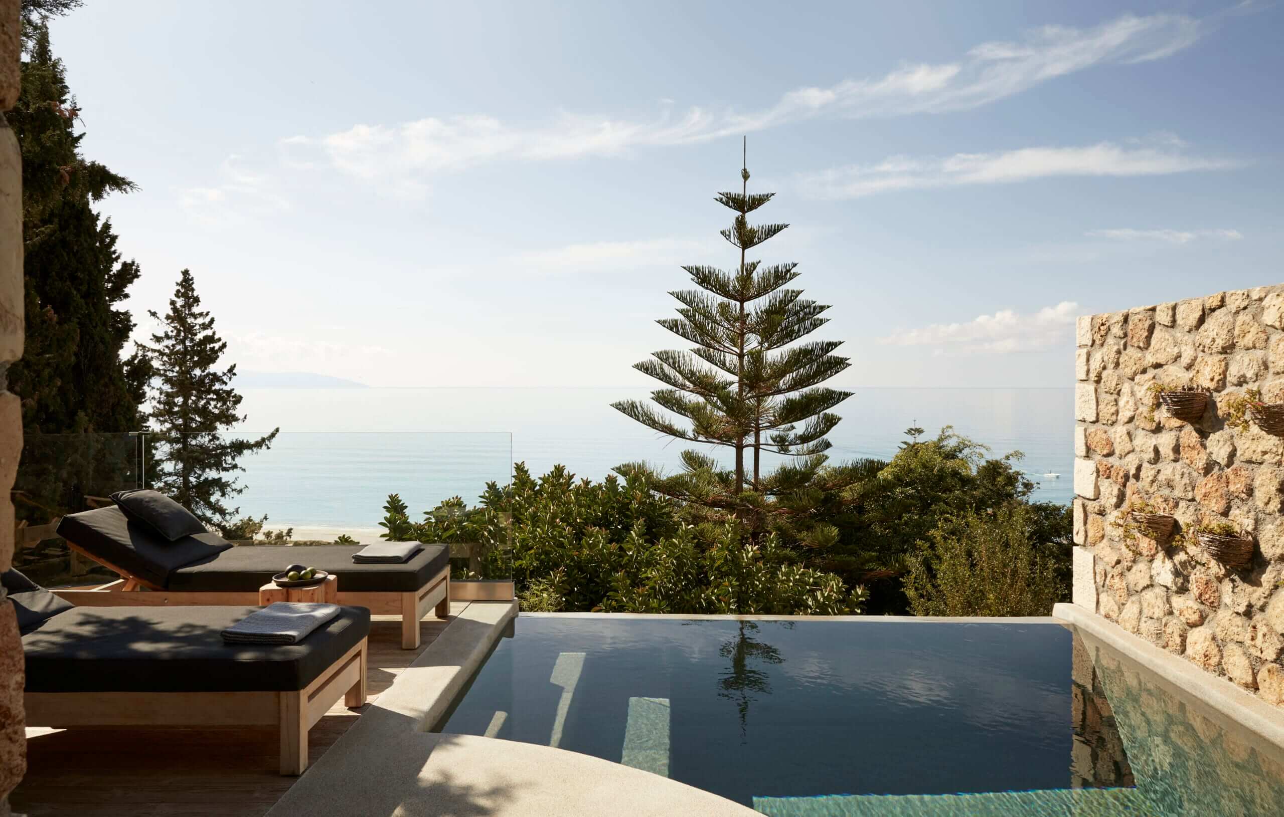 F Zeen Retreat, Europe — Infinity pool with stone walls overlooking Mediterranean, modernist lounge chairs, cypress trees, calm water reflecting sky.
