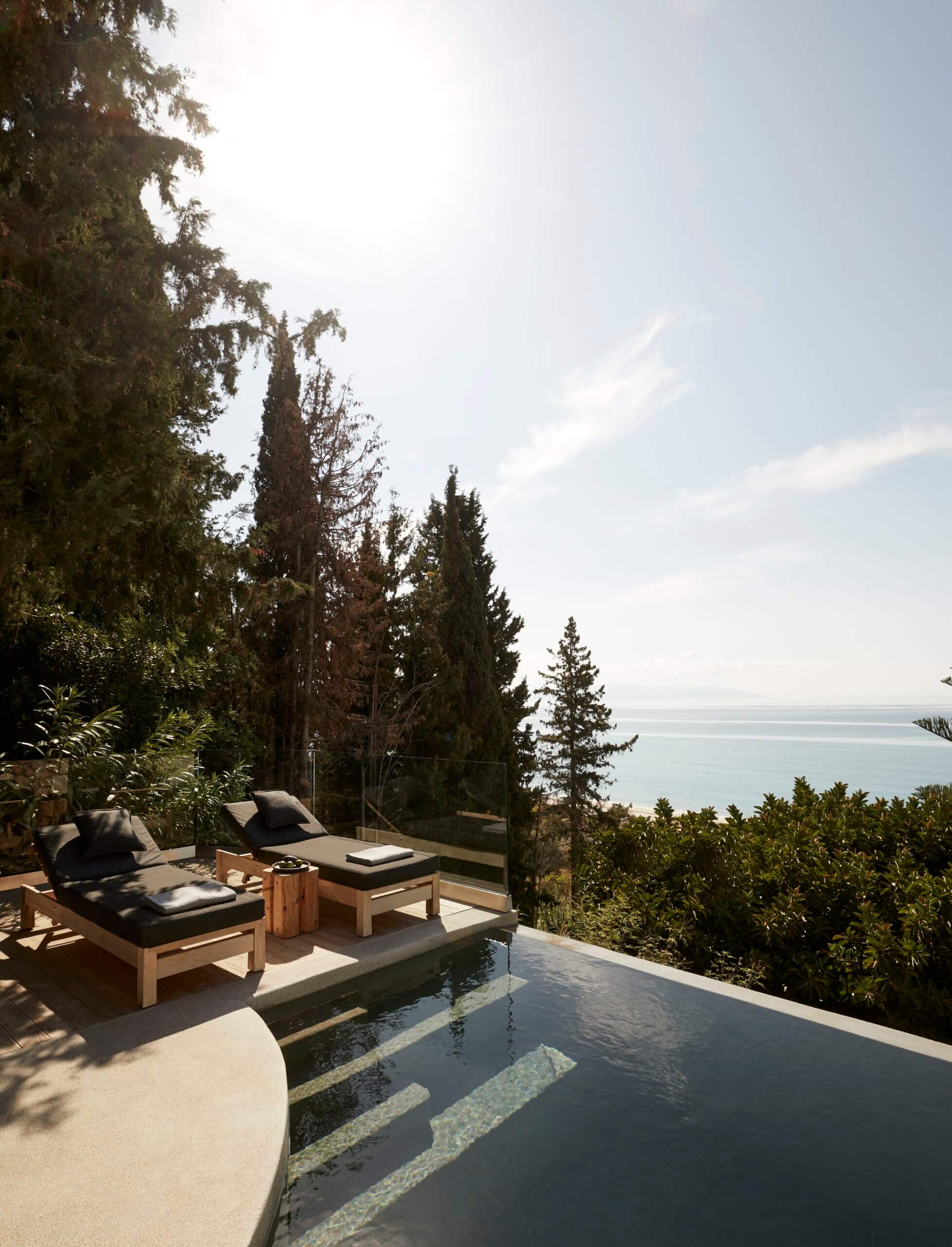 F Zeen Retreat, Europe — Infinity pool overlooking Mediterranean sea, cypress trees, lounge chairs with cushions, minimalist terrace architecture.