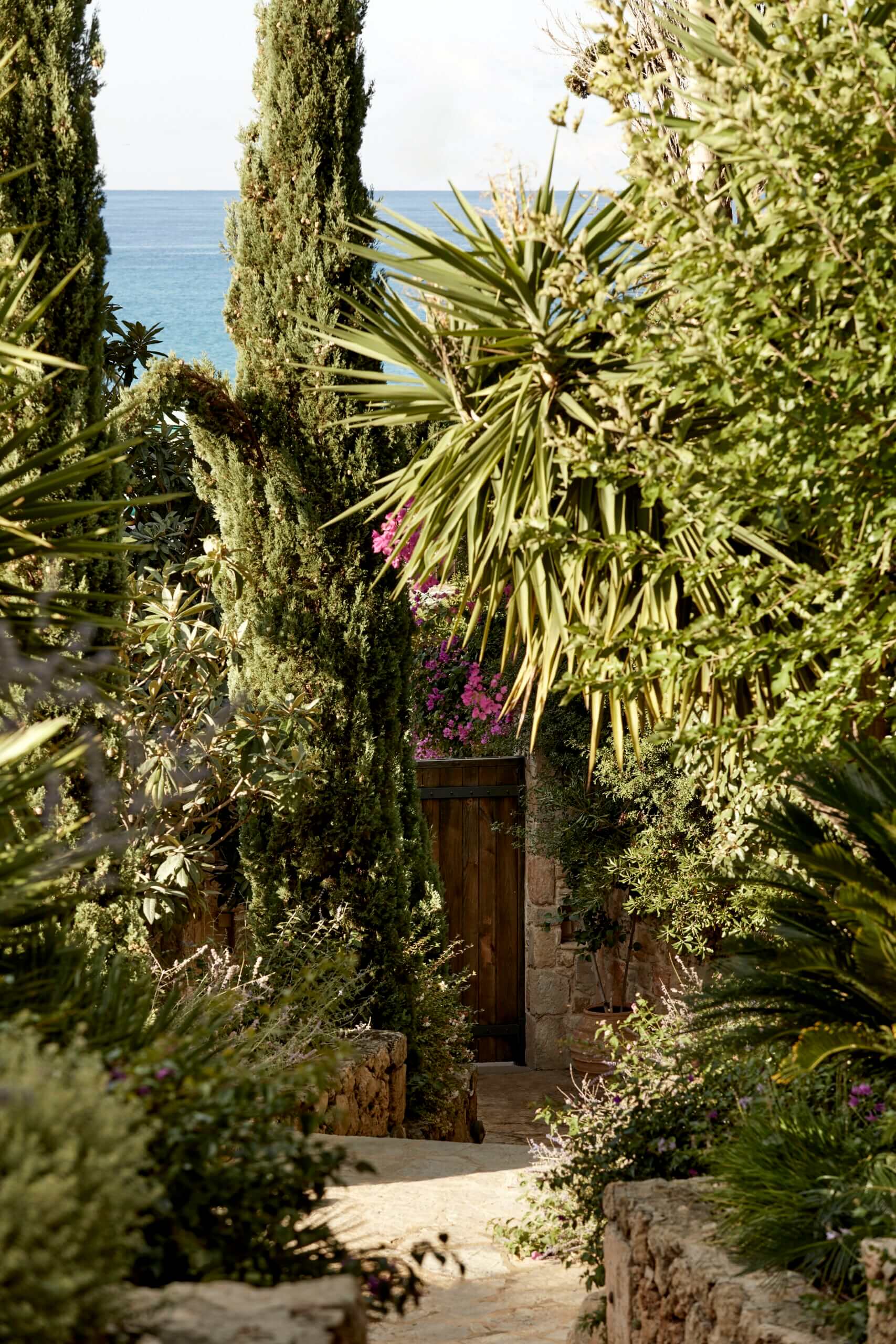 F Zeen Retreat, Europe — Mediterranean garden pathway with cypress trees, palm fronds, and magenta bougainvillea leading to wooden door with sea view beyond.