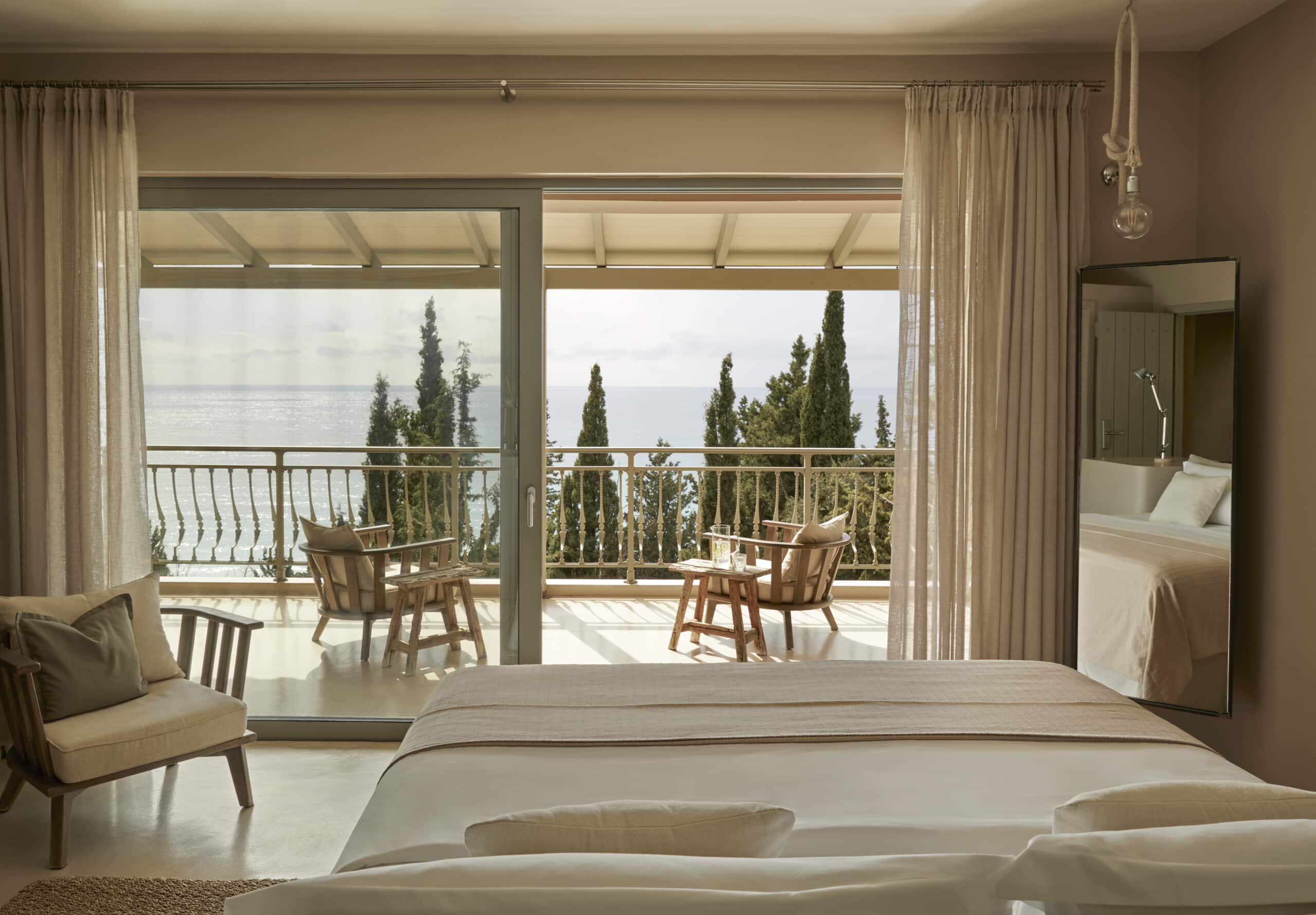 F Zeen Retreat, Europe — Modernist bedroom with floor-to-ceiling glass doors opening to sea-view terrace, cypress trees, mid-century wooden furniture, neutral linen, natural light.