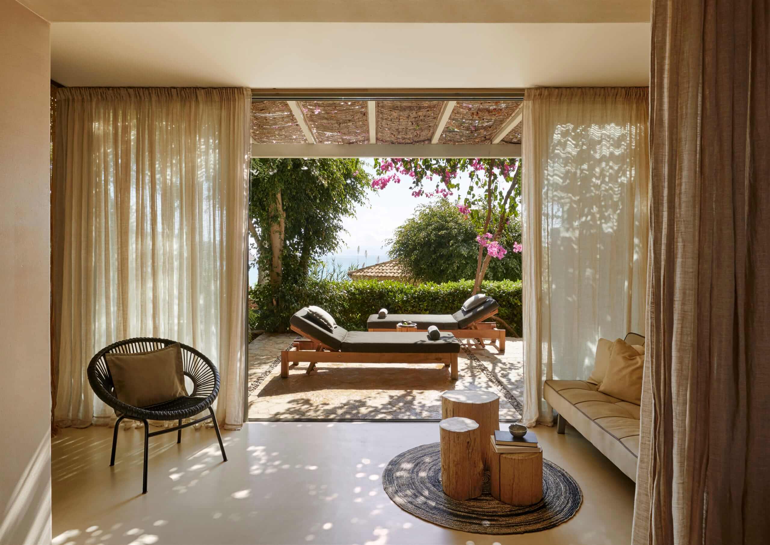 F Zeen Retreat, Europe — Sunlit interior opening to terrace with loungers, flowering bougainvillea, sheer curtains, and coastal views through open glass doors.