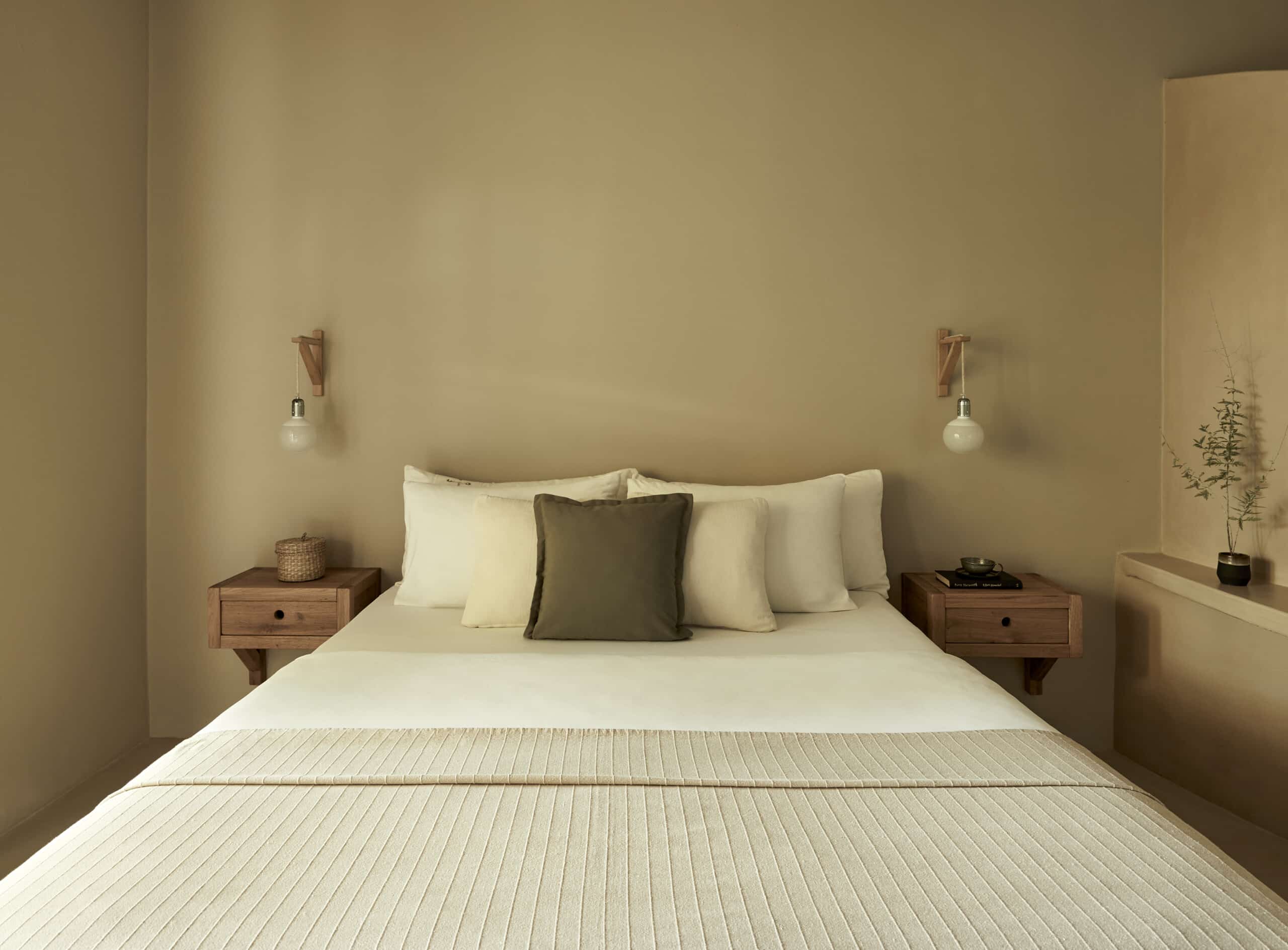 F Zeen Retreat, Europe — Minimalist bedroom with warm beige walls, walnut nightstands, cream bedding, pendant sconces, natural light, serene neutral aesthetic.