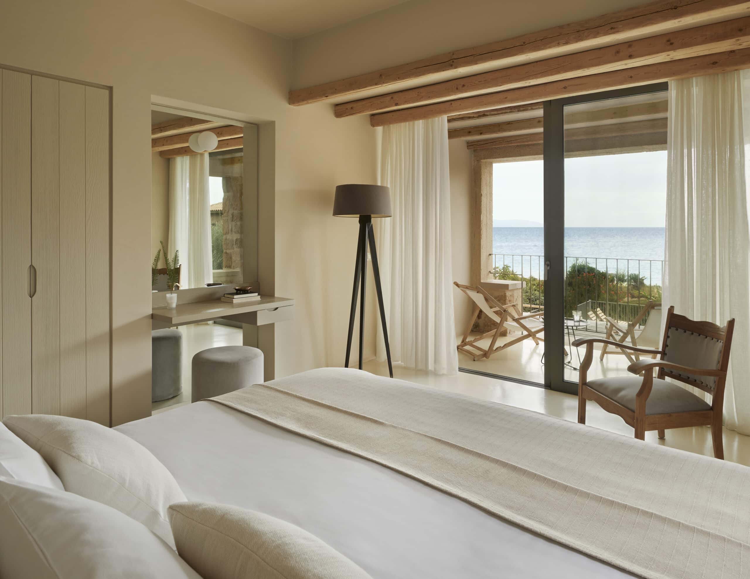 F Zeen Retreat, Europe — Minimalist beachfront bedroom with exposed wooden beams, cream linens, floor-to-ceiling glass doors opening to sea view terrace with lounge chairs.