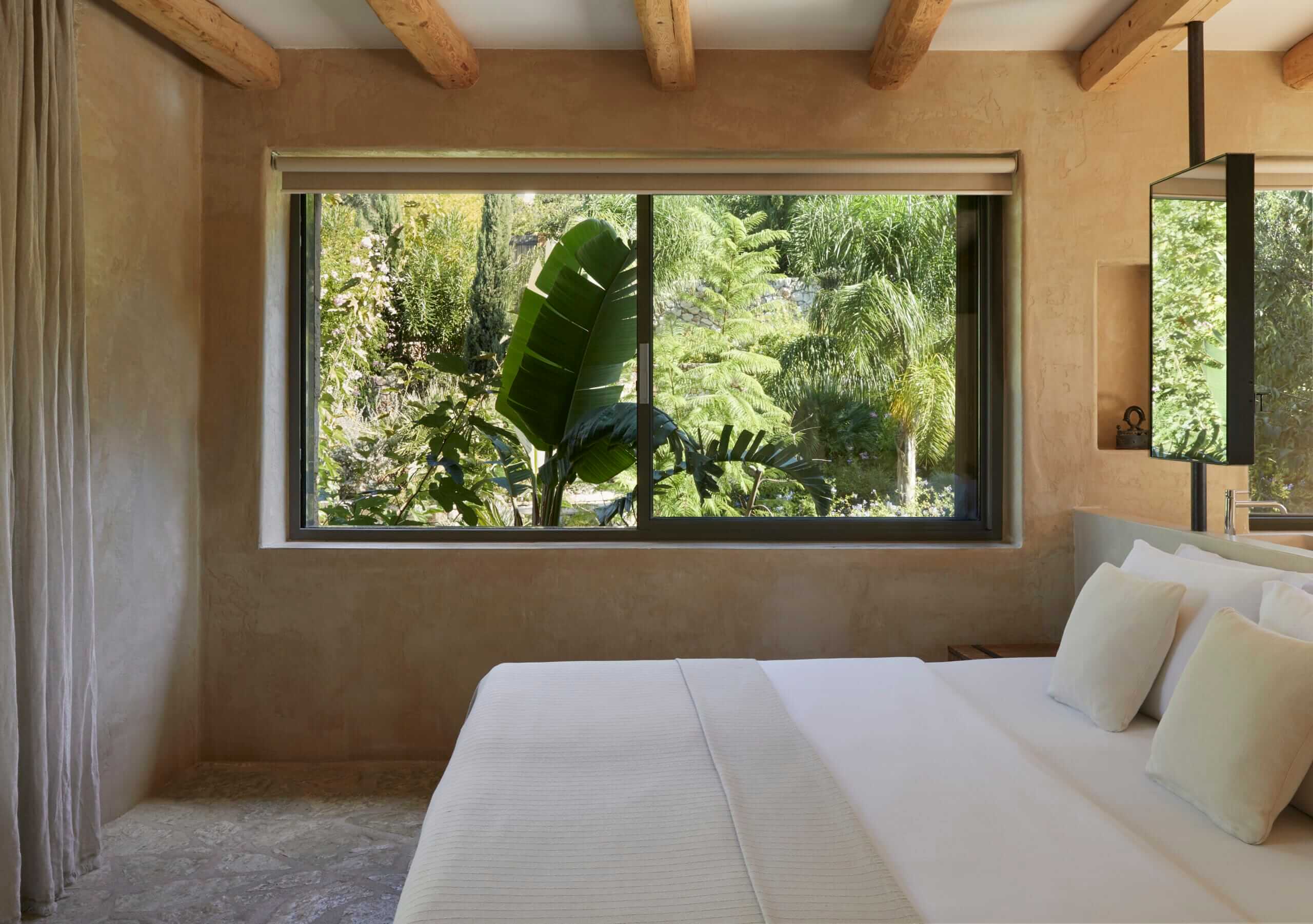 F Zeen Retreat, Europe — Minimalist bedroom with terracotta walls, exposed wooden beams, white linens, and expansive window framing lush tropical garden views.